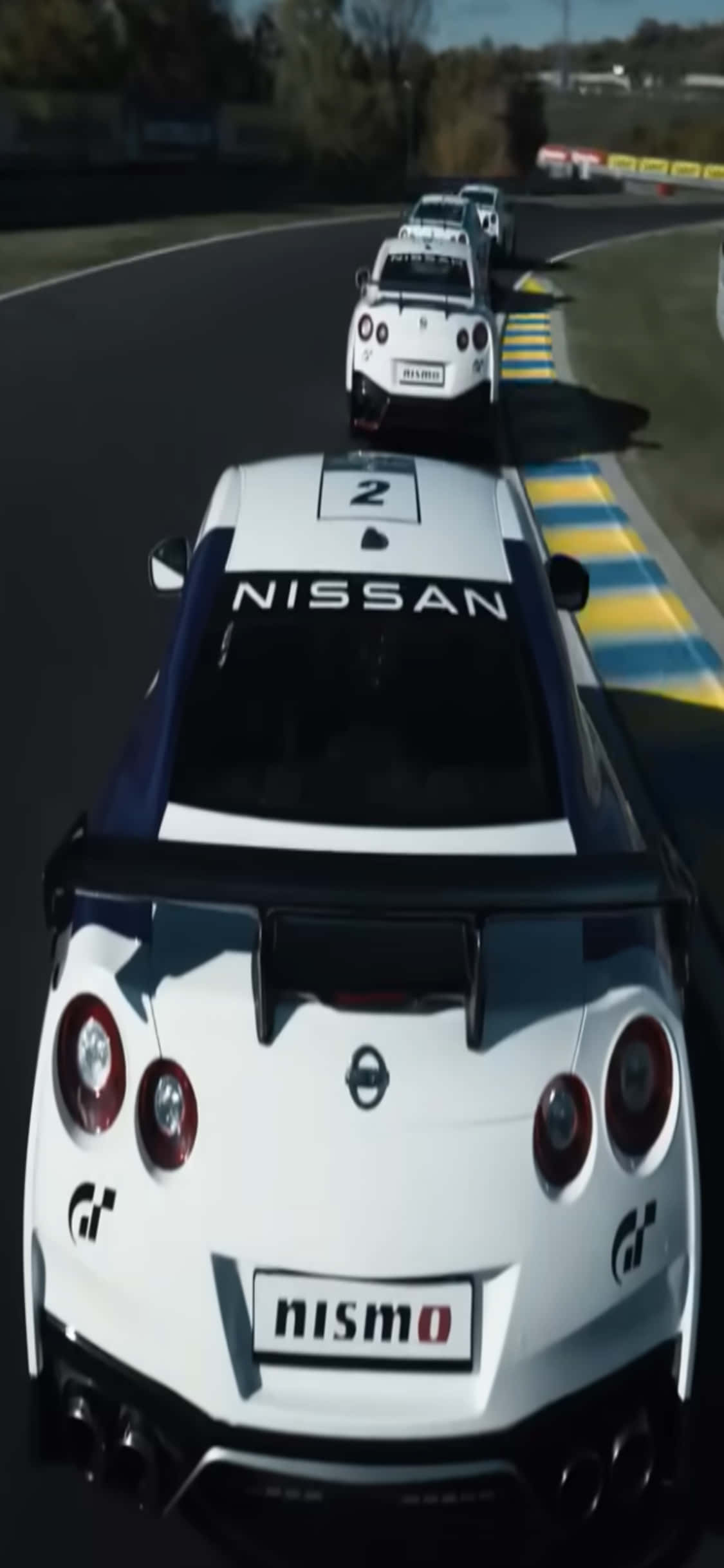 電玩遊戲 桌布 壁紙 — 一輛白色日產GT-R尼桑（Nismo）賽車，車號二，在明亮的日光下沿著一條彎曲的賽道奔馳，賽道邊緣有藍色和黃色的路緣石。