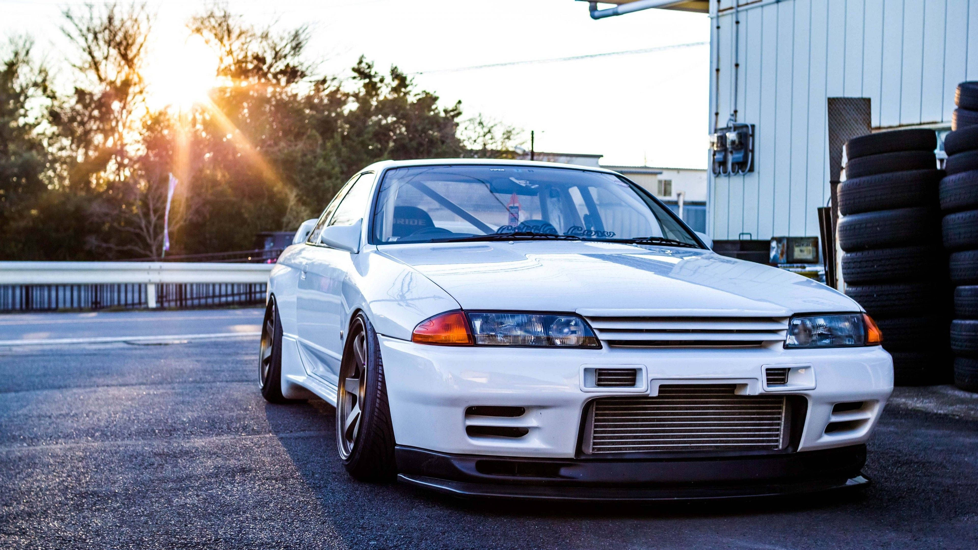 白色 桌布 壁紙 — 一輛白色日產Skyline GT-R停在柏油路上，樹後有陽光眩光，附近有堆疊的輪胎。