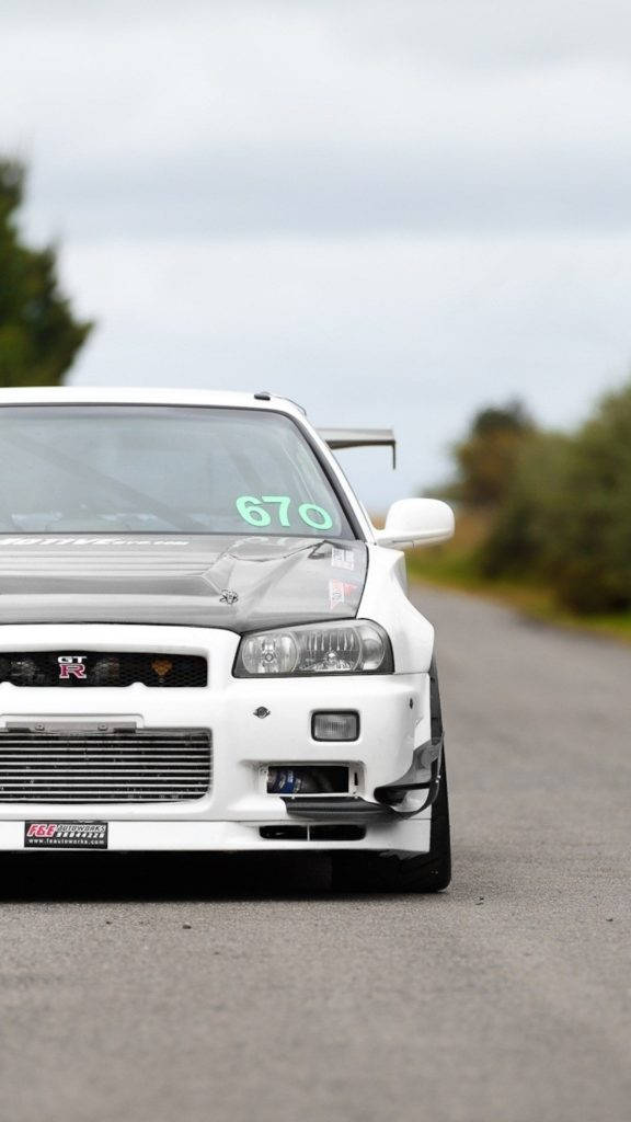 天際線 桌布 壁紙 — 一輛白色日產Skyline R34賽車，擋風玻璃上有670號碼，停泊在鄉村柏油路上。