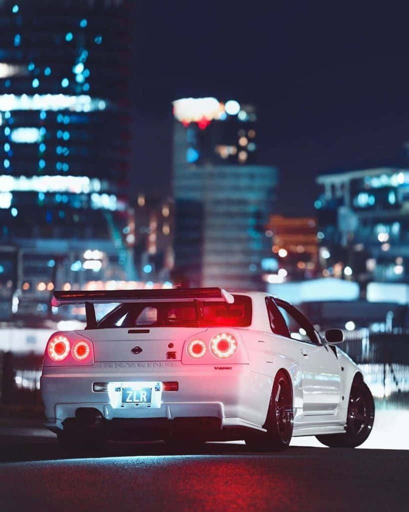 Buildings 桌布 壁紙 — 一輛白色日產Skyline R34跑車，帶有發光的紅色尾燈，停在夜晚潮濕的城市街道上，背景是模糊的霓虹燈。