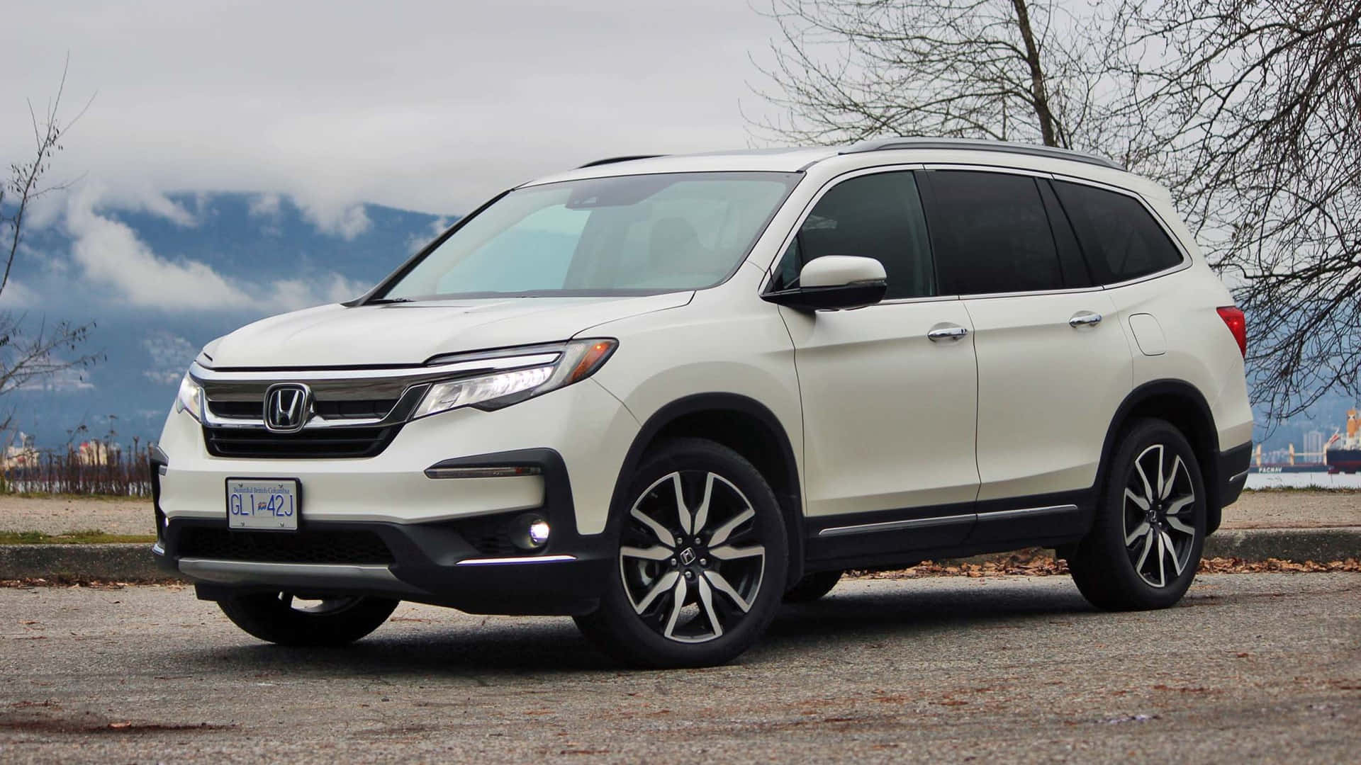 Honda Pilot 桌布 壁紙 — 一輛白色本田Pilot SUV停泊在碎石上，周圍有光禿禿的樹木和遠處的山脈，天空多雲。