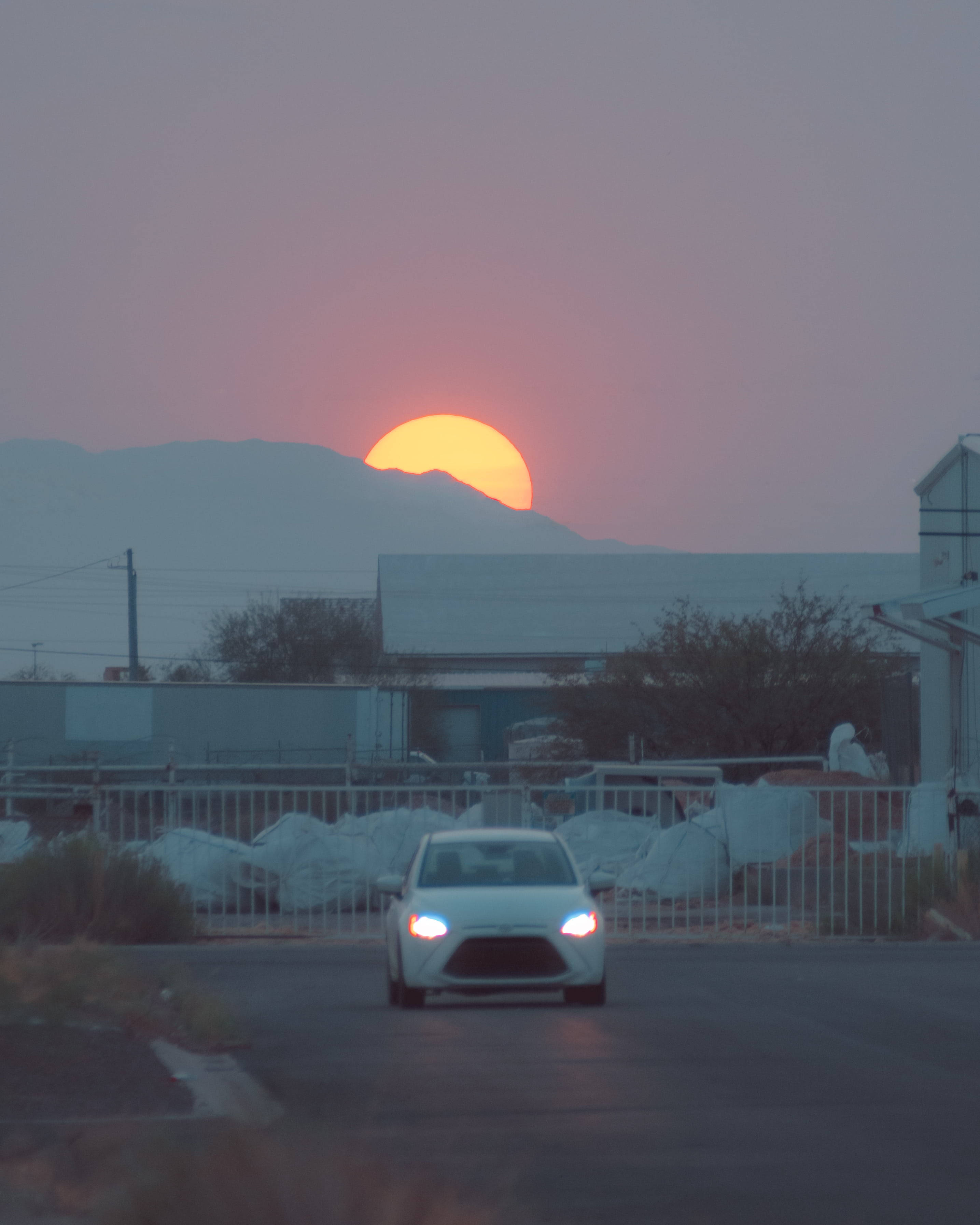 Aesthetic Sunset 桌布 壁紙 — 一輛白色汽車駛向觀眾，黃昏時分，一輪發光的橙色太陽落在遠處的山脈後面。