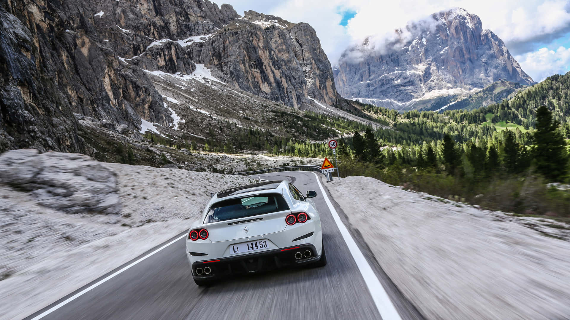 Ferrari Gtc4lusso 桌布 壁紙 — 一輛白色法拉利在部分多雲的天空下，沿著崎嶇山峰和綠色森林環繞的蜿蜒山路飛馳。