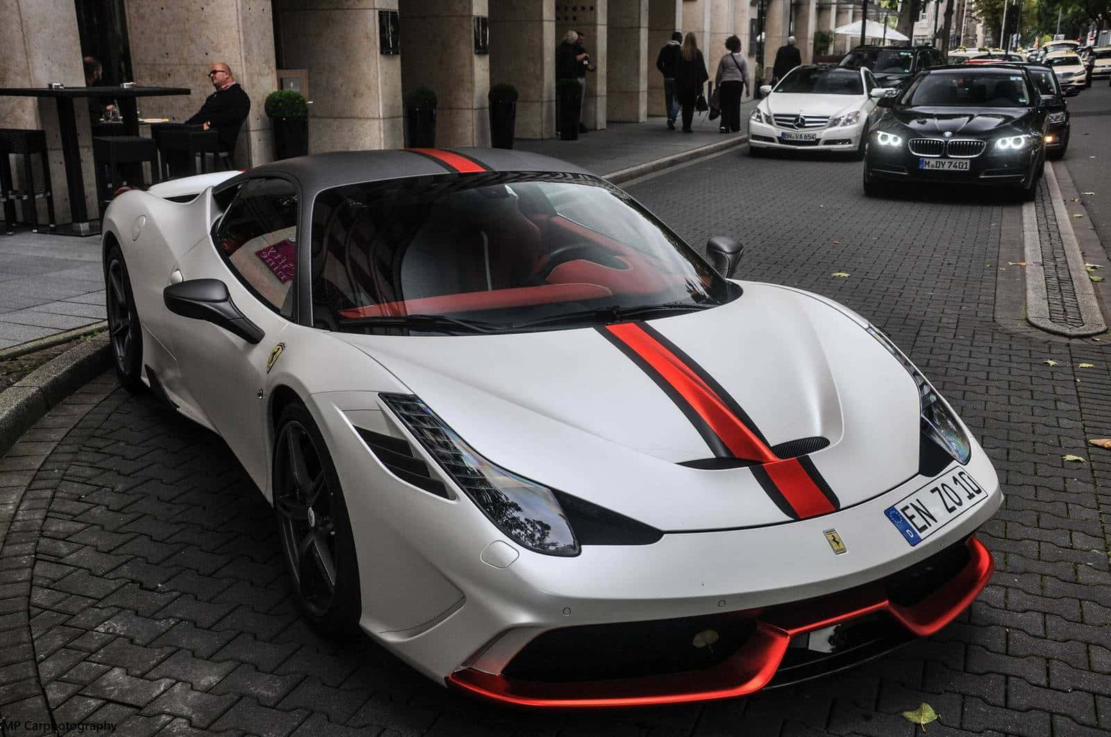 Ferrari 458 Speciale 桌布 壁紙 — 一輛白色法拉利帶有鮮明的紅色條紋，停在鵝卵石街道上，附近有咖啡館、行人和其它豪華車。