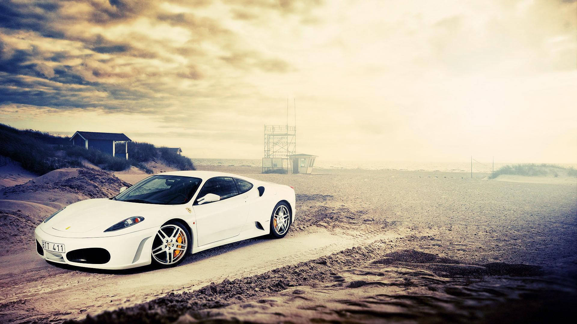 white ferrari 桌布 壁紙 — 一輛白色法拉利跑車停泊在沙灘上，天空多雲，有沙丘和遠處的建築物。