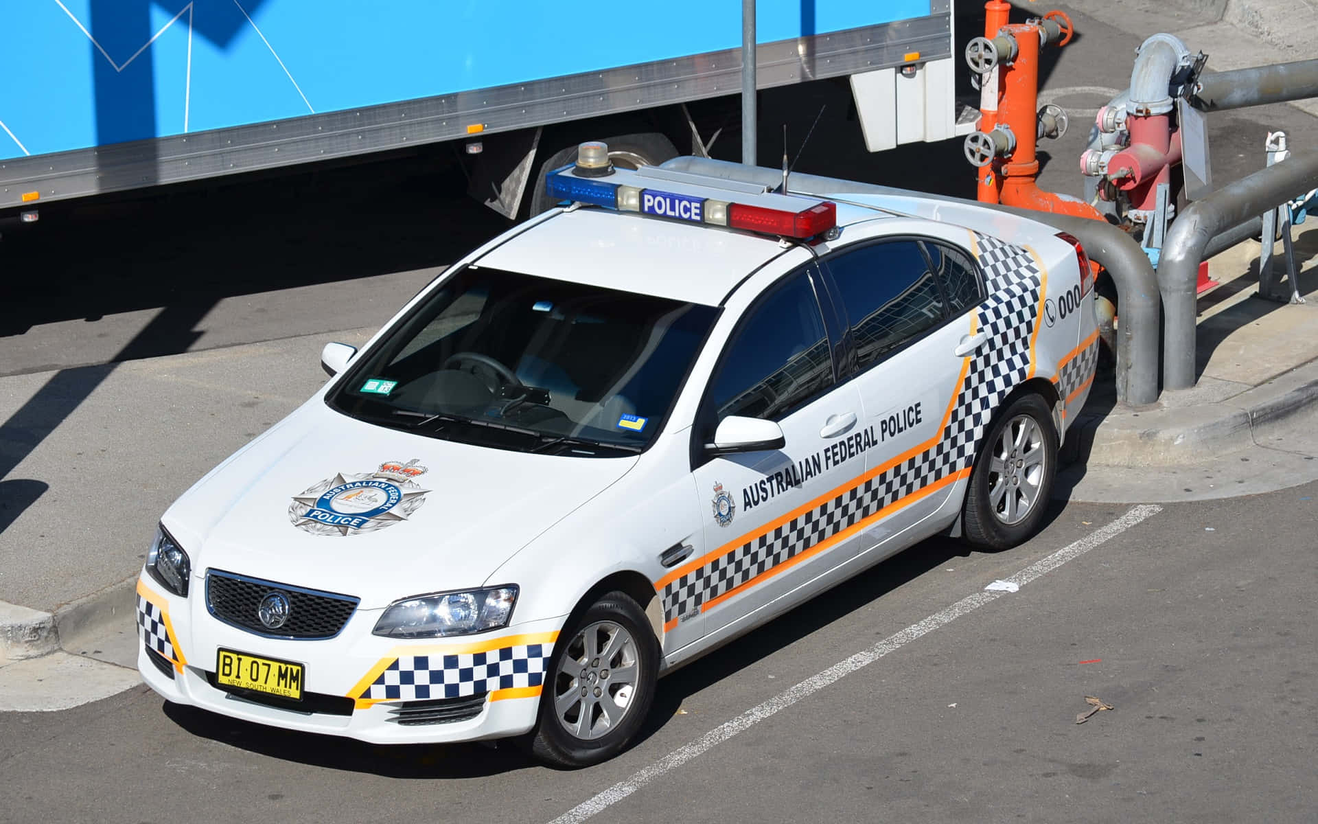 Law Enforcement 桌布 壁紙 — 一輛白色澳洲聯邦警車，帶有格子和橙色條紋，在明亮日光下停在一輛藍色卡車旁邊。