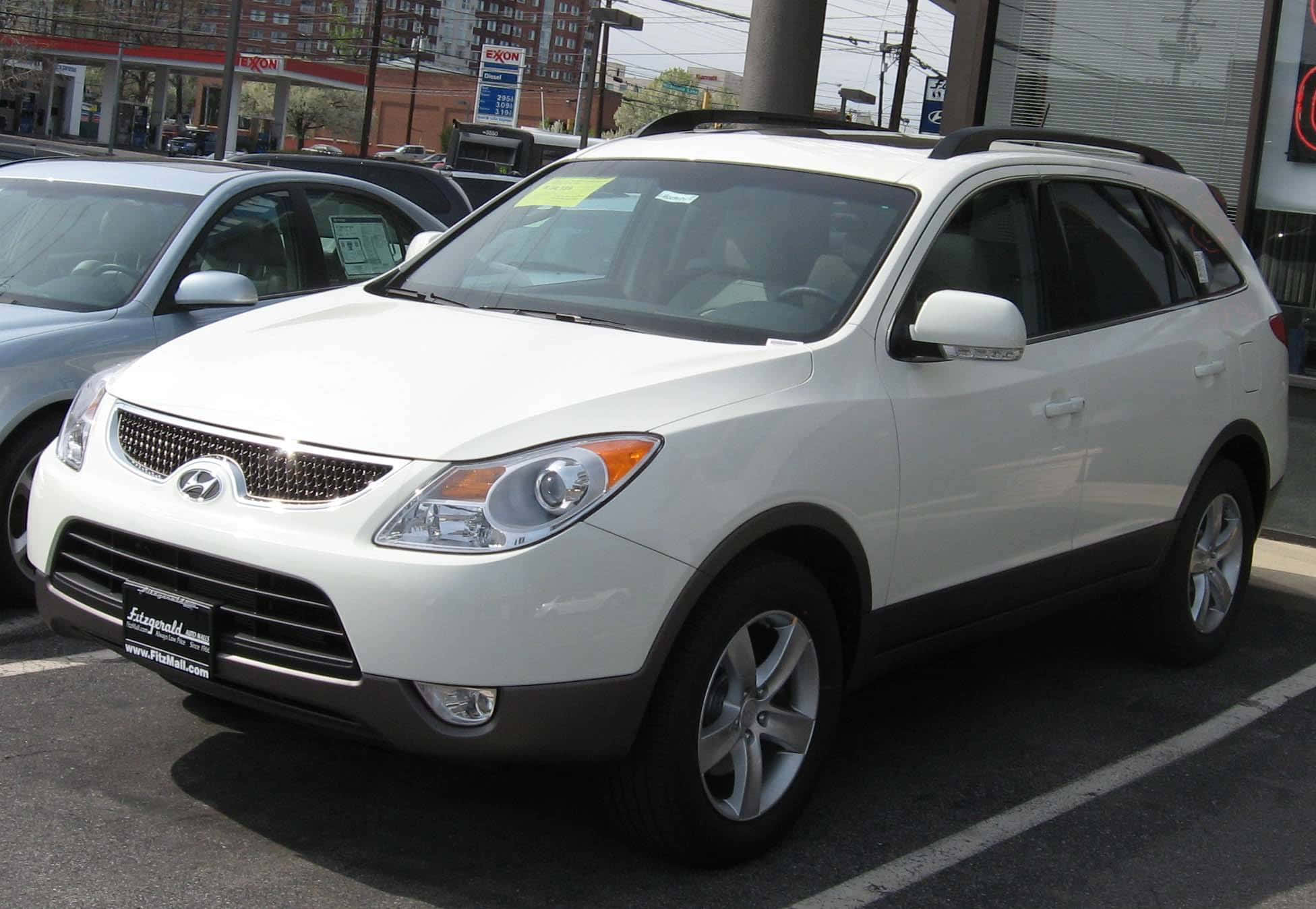 Hyundai Veracruz 桌布 壁紙 — 一輛白色現代Tucson SUV停放在經銷商停車場，在明亮的日光下，背景有其他汽車和建築物。