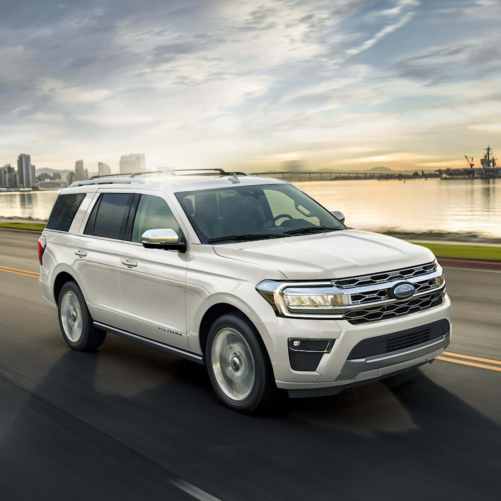 福特 桌布 壁紙 — 一輛白色福特 Expedition SUV 沿著海濱道路行駛，背景是城市天際線和船隻，正值黃金時刻。