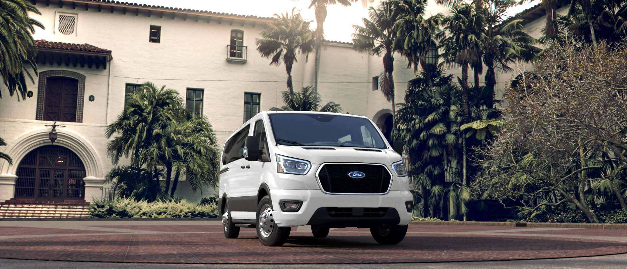 Ford Transit 桌布 壁紙 — 一輛白色福特 Transit 客貨車停放在磚砌車道上，前方是西班牙風格建築，周圍環繞著棕櫚樹。