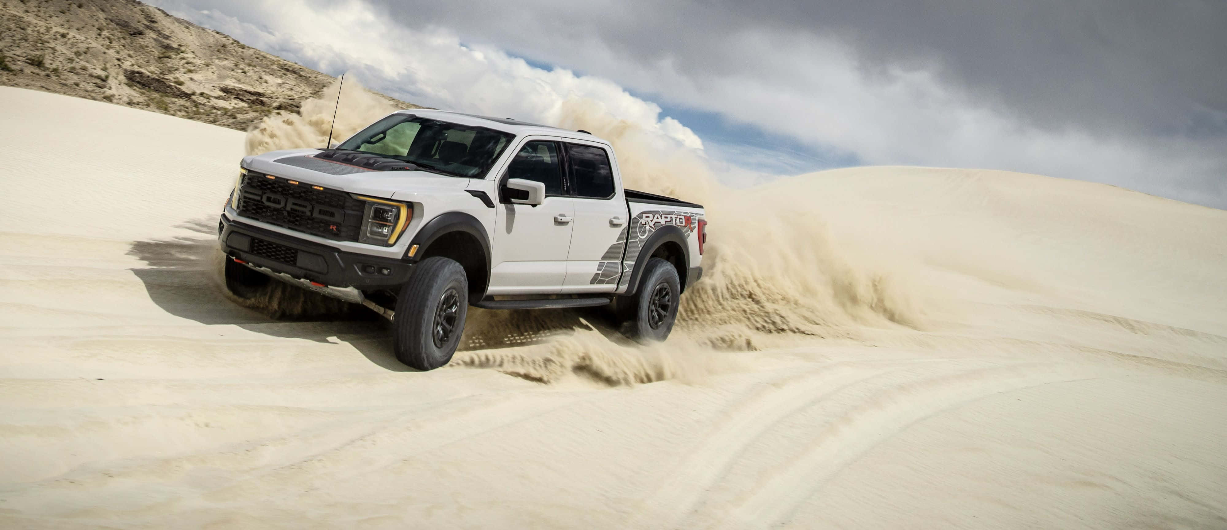 Ford F 150 桌布 壁紙 — 一輛白色福特F-150 Raptor卡車在多雲的天空下駛過廣闊的沙漠沙丘，揚起沙塵。