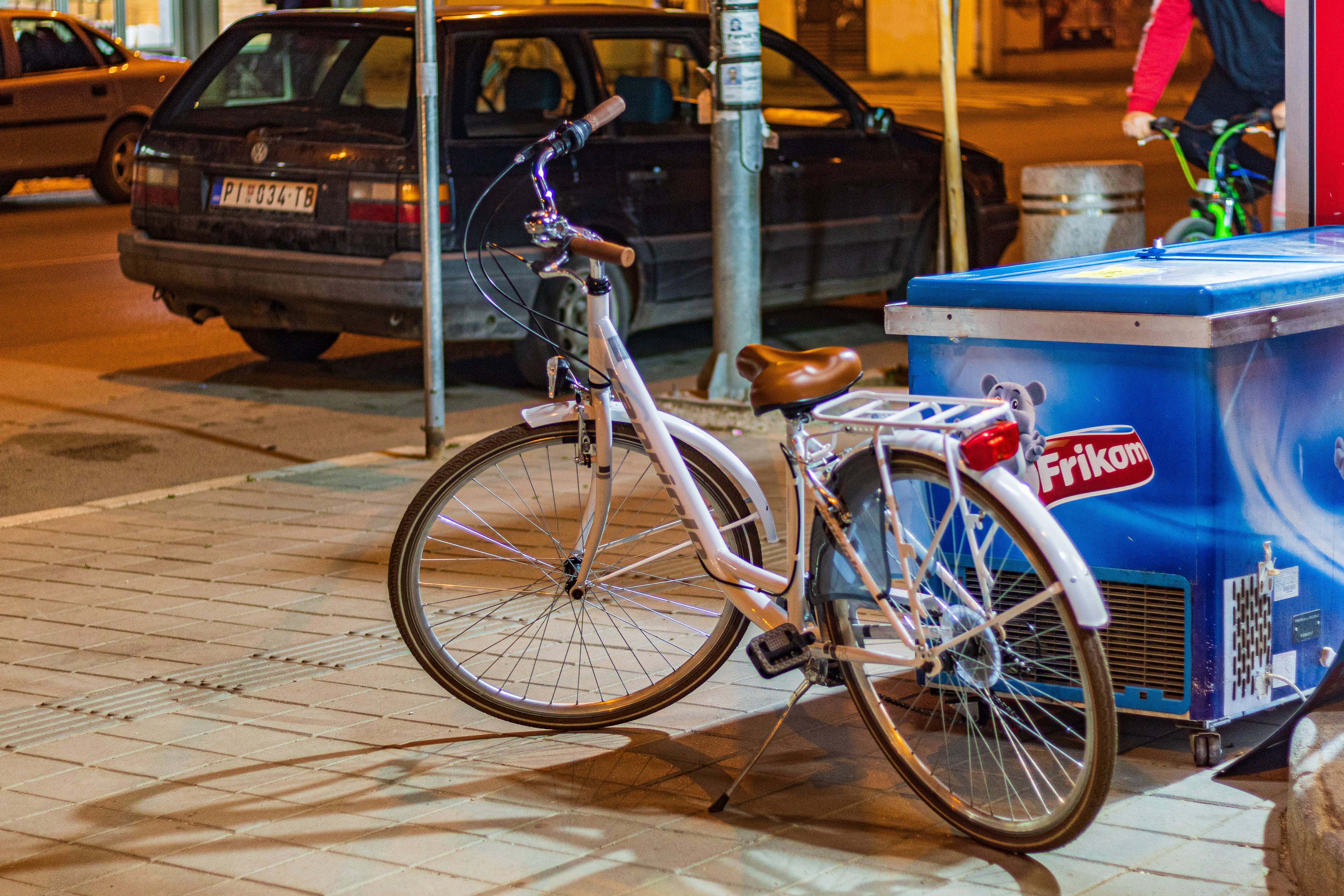 Serbia 桌布 壁紙 — 一輛白色腳踏車配棕色座椅，夜晚停泊在瓷磚人行道上，旁邊有一輛藍色冰淇淋車，在溫暖街燈下。