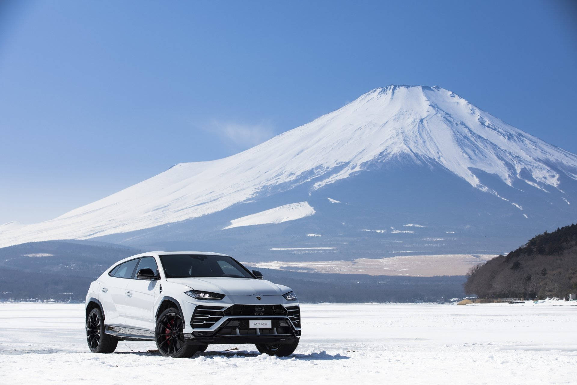 lambo 桌布 壁紙 — 一輛白色藍寶堅尼 Urus 停在結冰的湖面上，背後是白雪覆蓋的富士山，在清澈的藍天下巍然聳立。