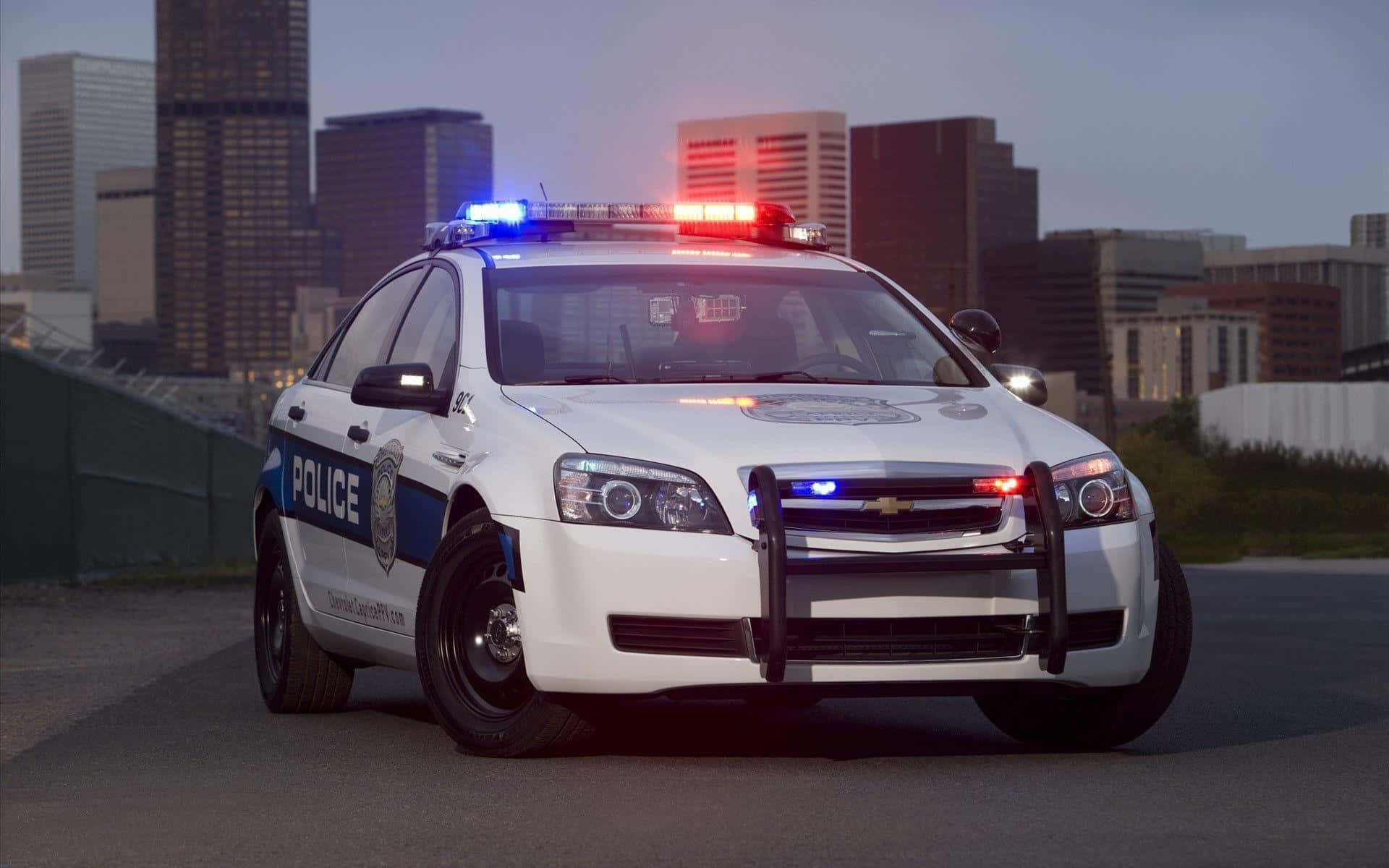 Police Car 桌布 壁紙 — 一輛白色警車在黃昏時分停泊在城市的街道上，紅藍燈閃爍，背景是高樓大廈。
