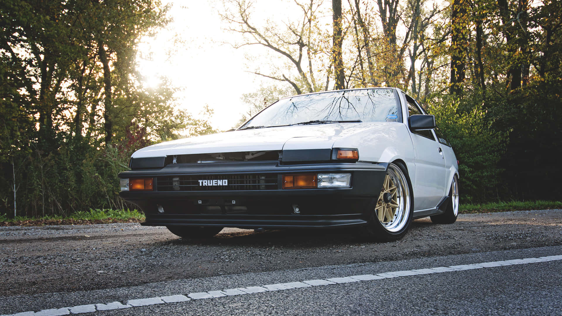 白色豐田 Trueno AE86 配金色輪圈，停泊在日落時分的森林道路上。 — Toyota Supra Mk4 壁紙