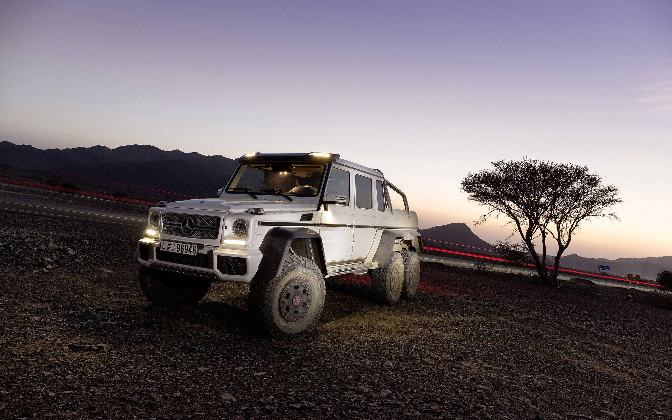Mercedes 桌布 壁紙 — 一輛白色賓士 G63 AMG 6x6 在黃昏時停在碎石上，車流燈光劃過，孤樹襯托紫色山巒。