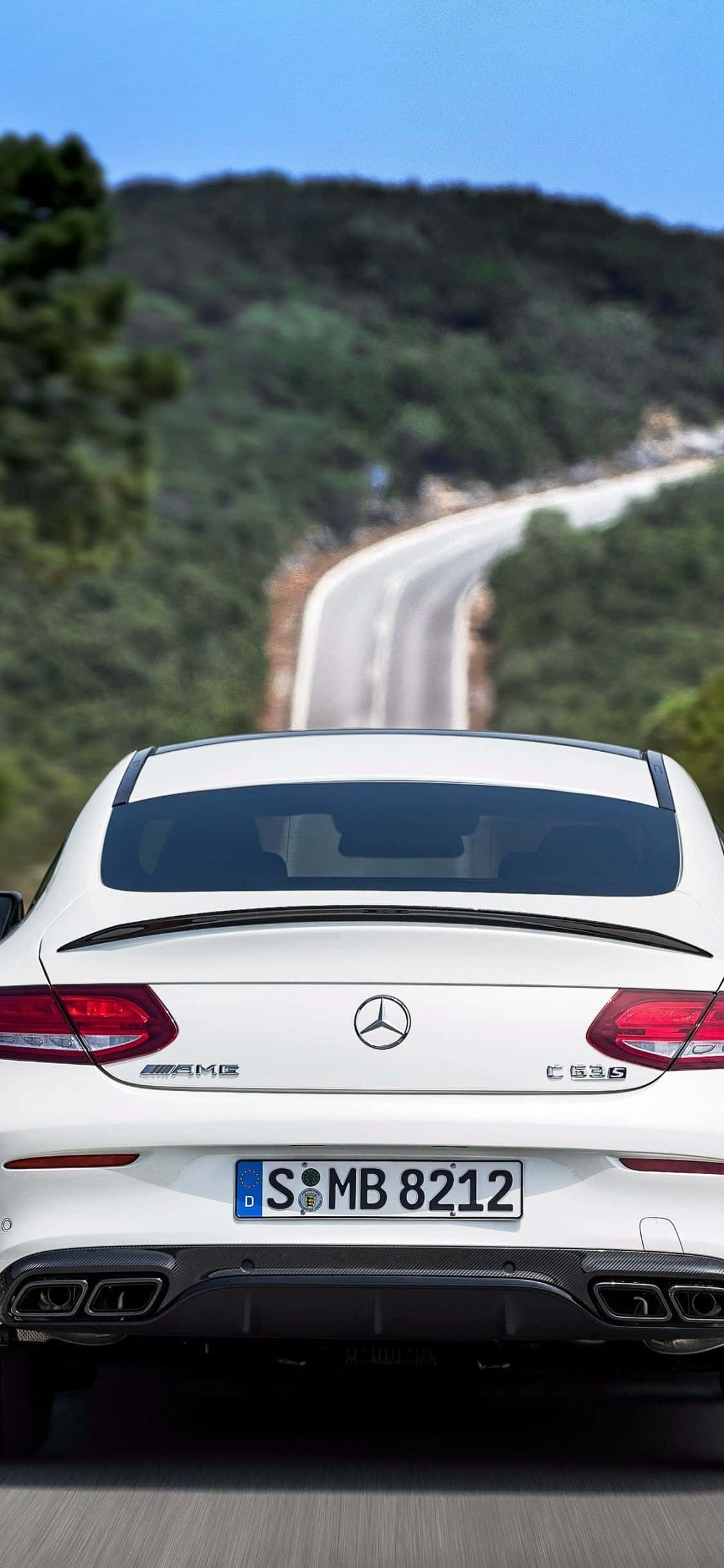 Mercedes 桌布 壁紙 — 一輛白色賓士AMG C63 S雙門跑車從後方觀看，在晴朗藍天下駛過蜿蜒山路，穿越翠綠山丘。