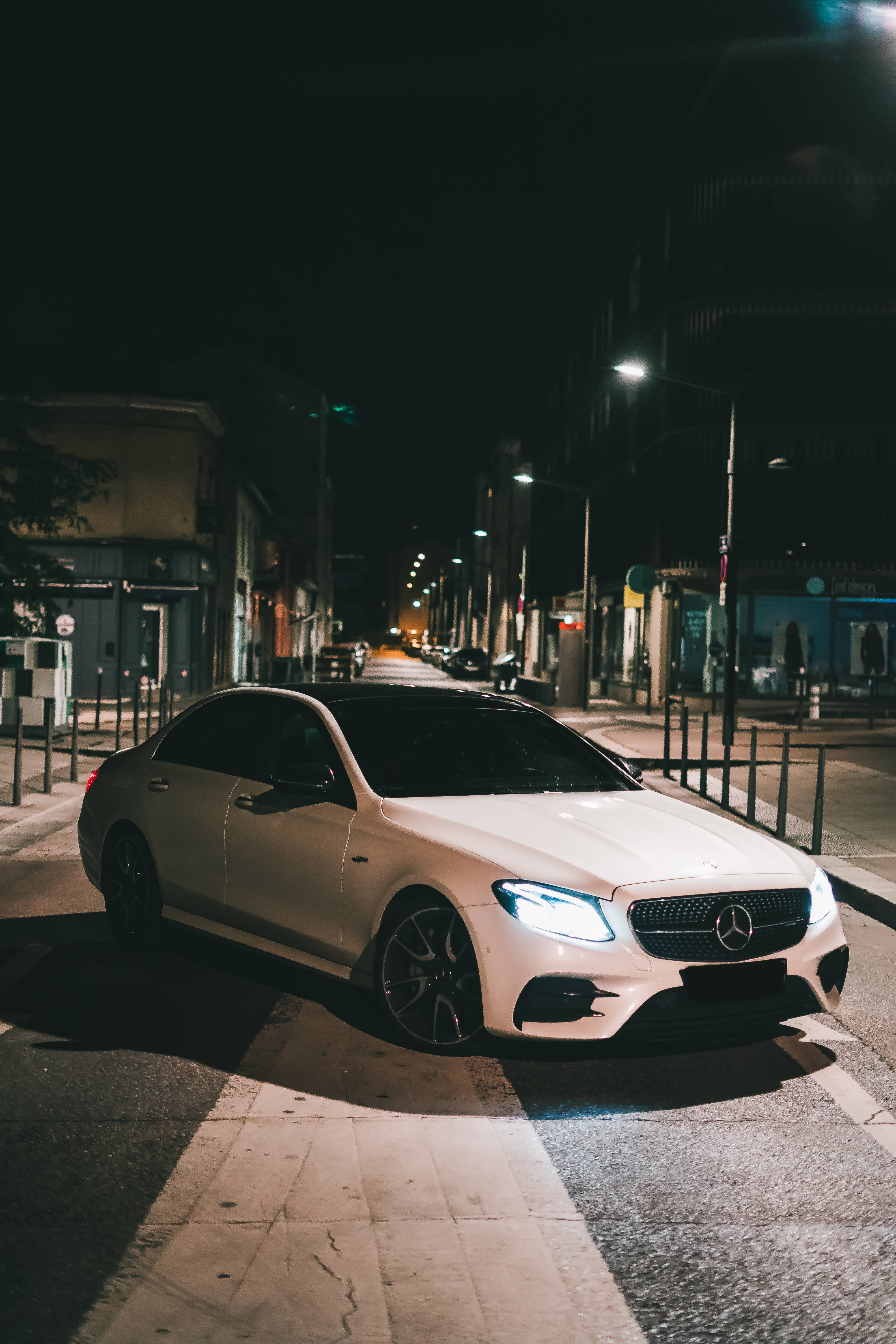 amg iPhone 桌布 壁紙 — 一輛白色賓士轎車在路燈下閃耀，夜晚停放在寧靜的城市街道上。