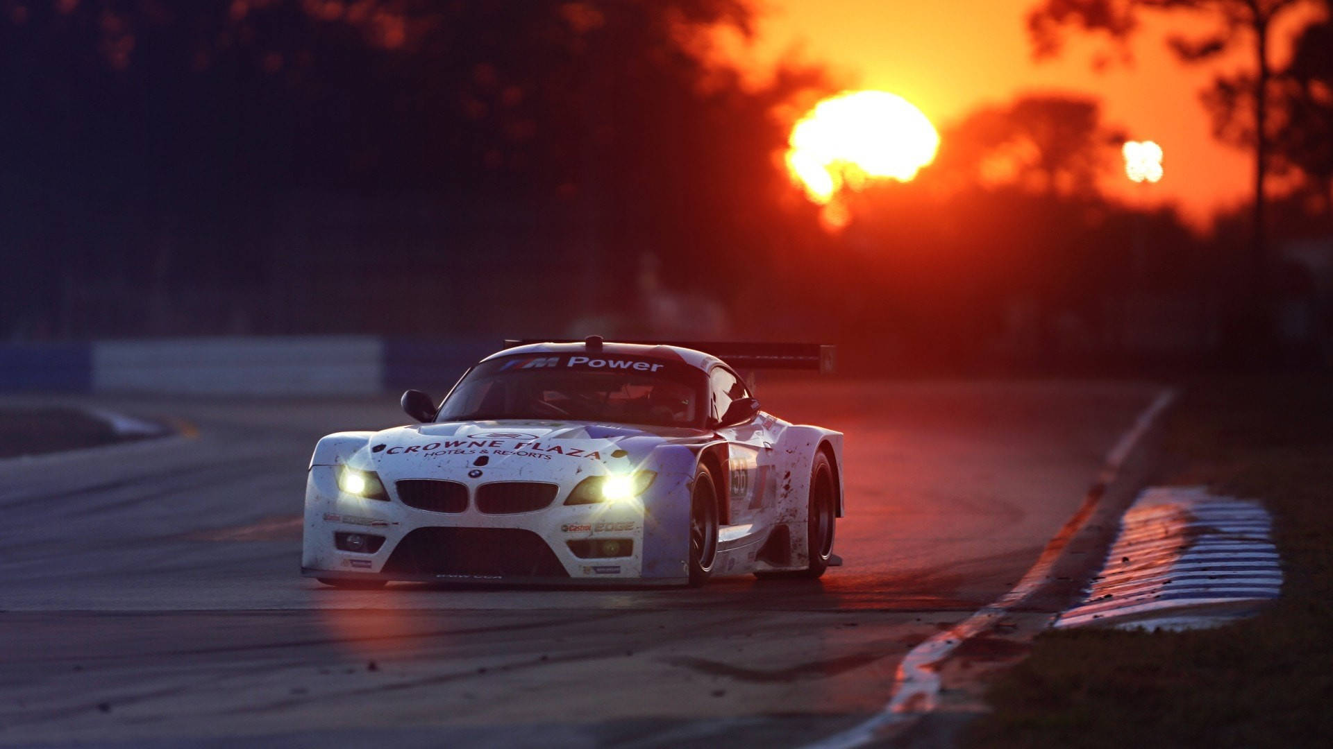 Auto Racing 桌布 壁紙 — 一輛白色賽車亮著車頭燈，在濕滑的賽道上疾馳，身後是熾熱的夕陽。
