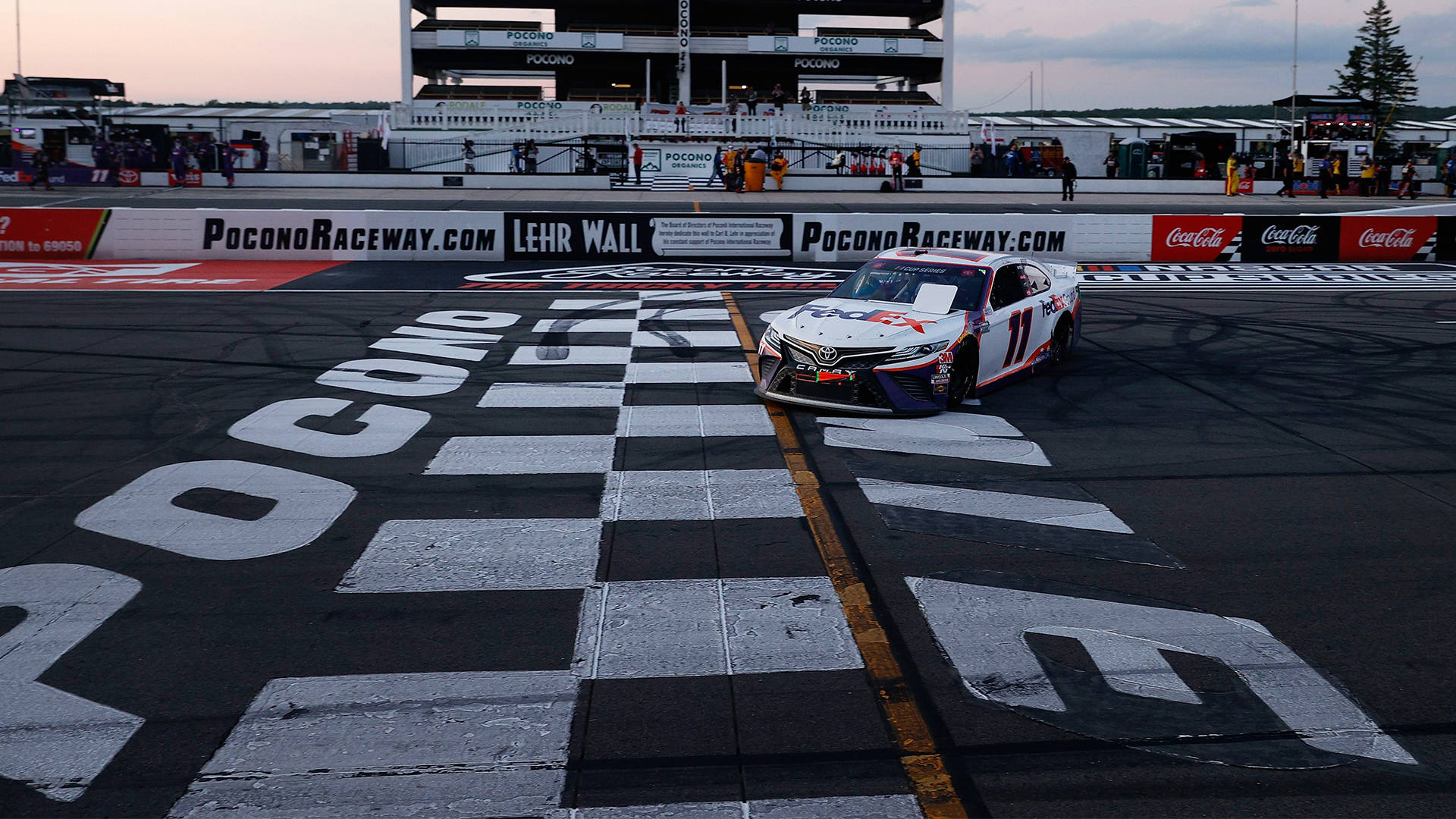 Denny Hamlin 桌布 壁紙 — 一輛白色賽車在黃昏時分越過波科諾賽道的方格旗終點線，背景有觀眾和維修站人員。