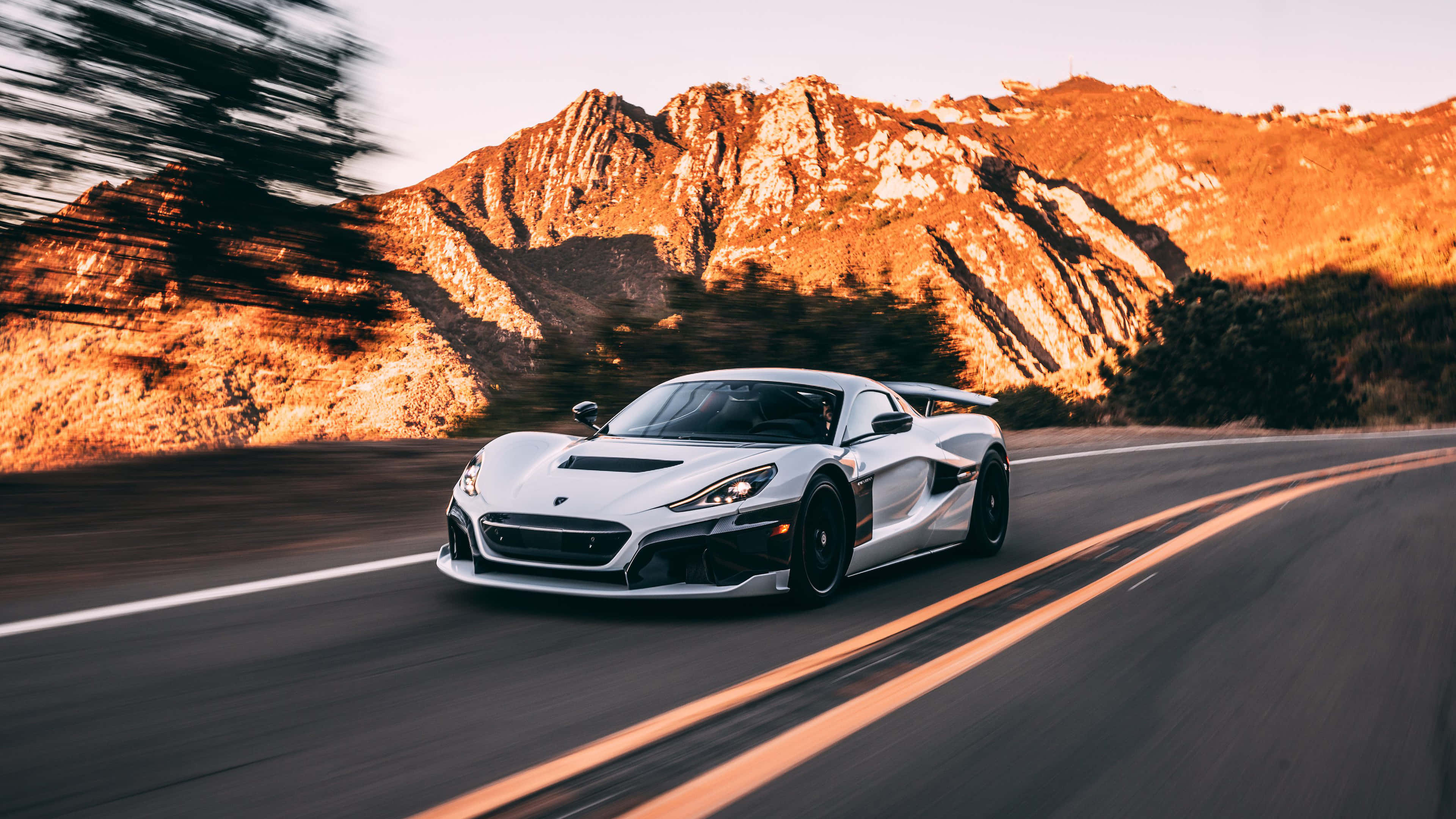 Rimac 桌布 壁紙 — 一輛白色超跑在日落時分沿著蜿蜒的山路疾馳，帶有模糊動感和金色光線。