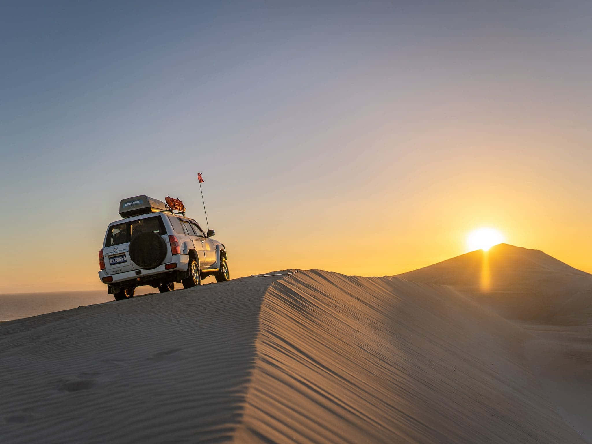 Sand Dunes 桌布 壁紙 — 一輛白色越野車停在沙丘頂端，夕陽的金色光芒在沙漠中投下長長的影子。
