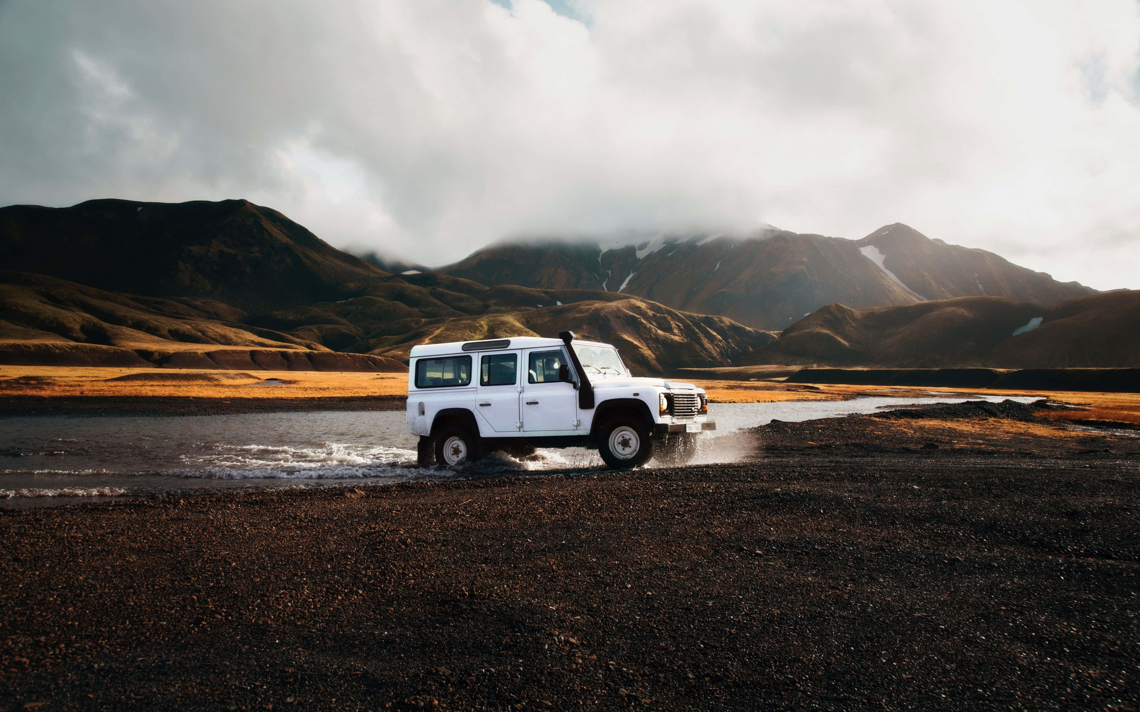 Best Travel 桌布 壁紙 — 一輛白色越野車在淺溪中濺起水花，背景是崎嶇的山脈和陰雲密布的天空。