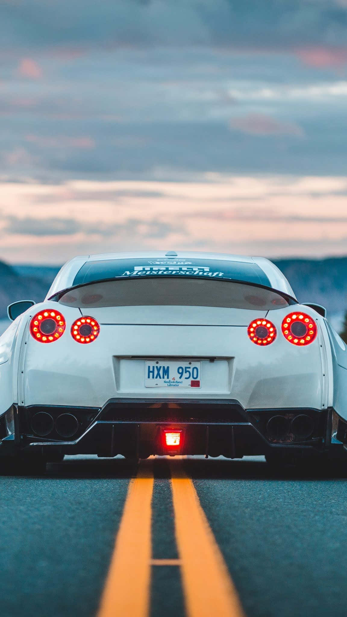 R33 GT-R 桌布 壁紙 — 一輛白色跑車亮著紅色尾燈，在黃昏時分行駛在道路上，背景是山脈。
