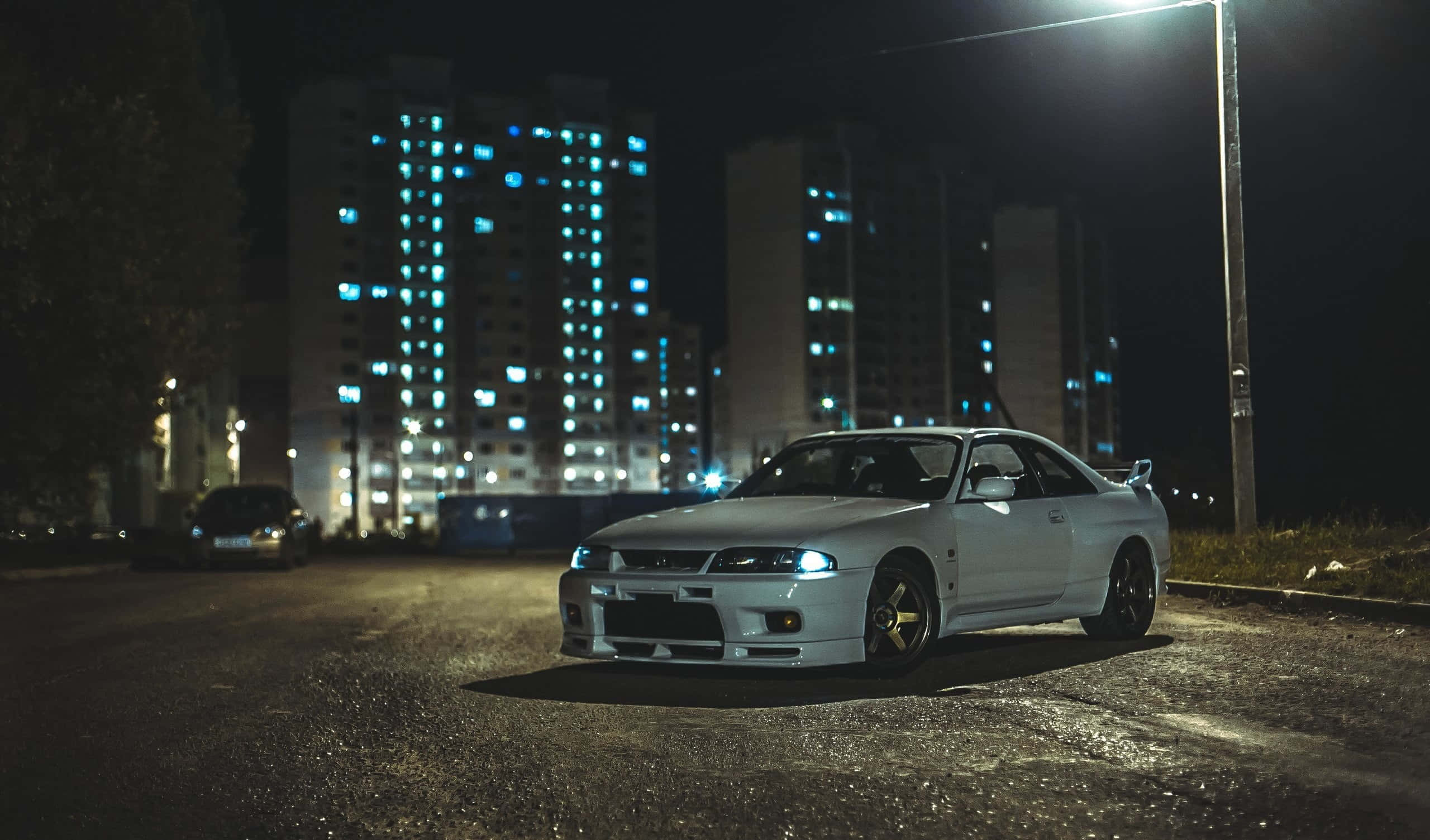 R33 GT-R 桌布 壁紙 — 一輛白色跑車在夜間濕潤的城市街道上，在街燈下閃閃發光，背景是發光的公寓大樓。