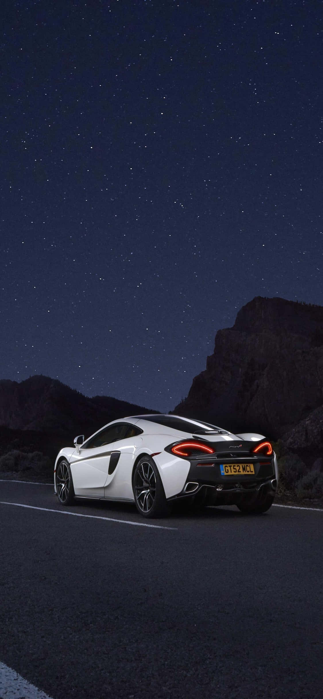 iPhone Xs Mclaren 720s 桌布 壁紙 — 一輛白色跑車在星空下的山路上發光，周圍是黑暗的岩石峭壁。