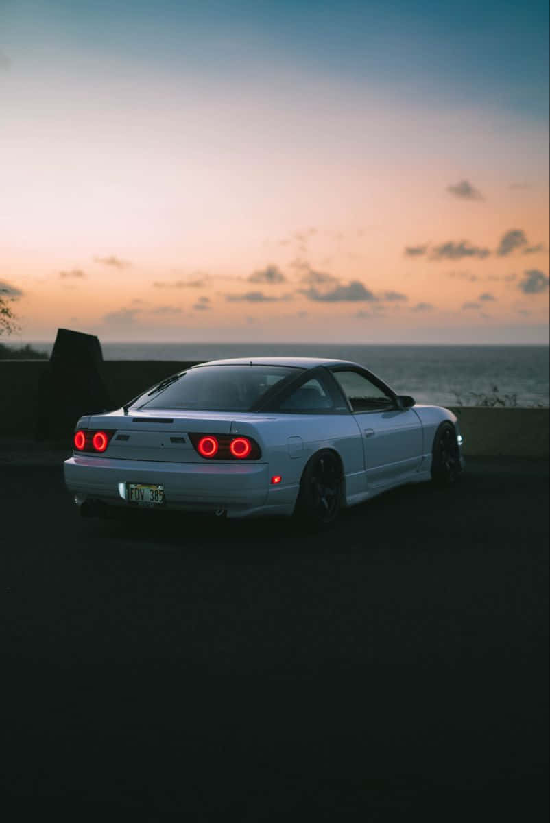 Nissan 180sx 桌布 壁紙 — 一輛白色跑車帶有發光的紅色尾燈，停在懸崖上，俯瞰日落時分的海洋。