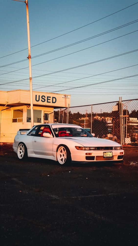 Nissan Silvia S13 桌布 壁紙 — 一輛白色跑車搭配紅色內裝，在日落時分停放在藍天白雲下的 'USED' 二手車場前。
