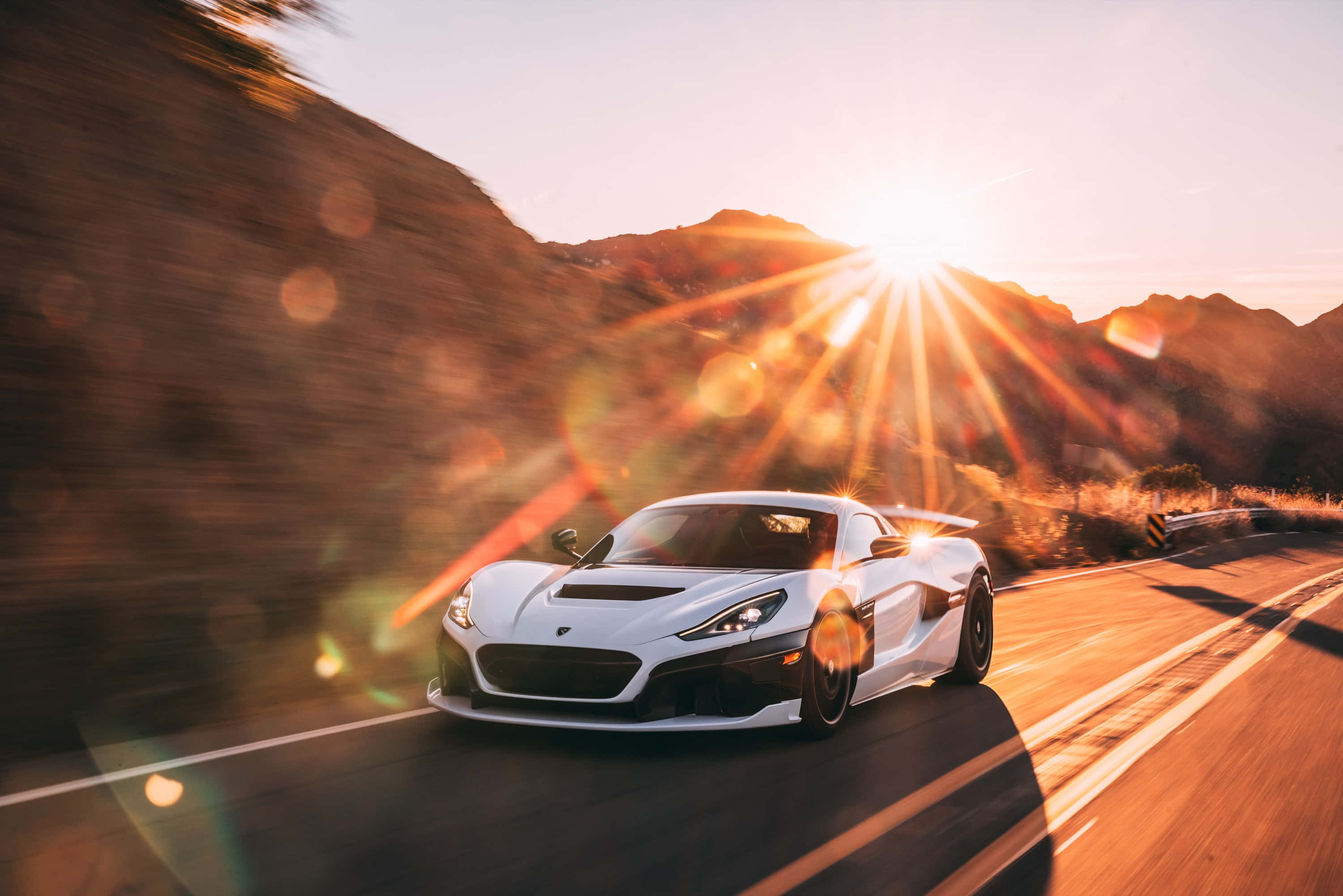 Rimac 桌布 壁紙 — 一輛白色跑車沿著蜿蜒的山路疾馳，太陽從後方山峰間迸發，形成鏡頭光暈。