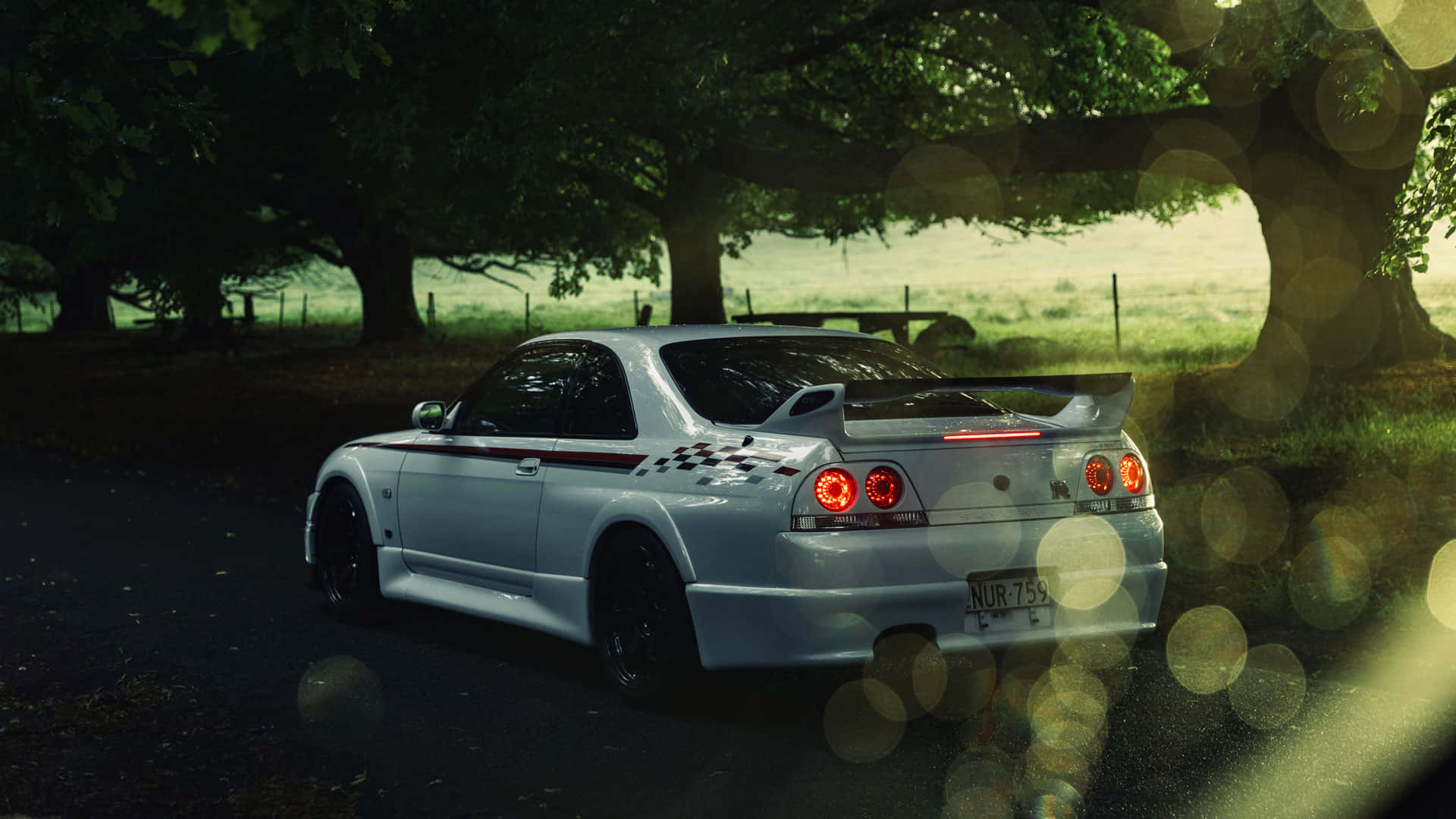 R33 GT-R 桌布 壁紙 — 一輛白色跑車配紅色尾燈，停放在枝葉茂密的樹下，前景有金色散景燈光。