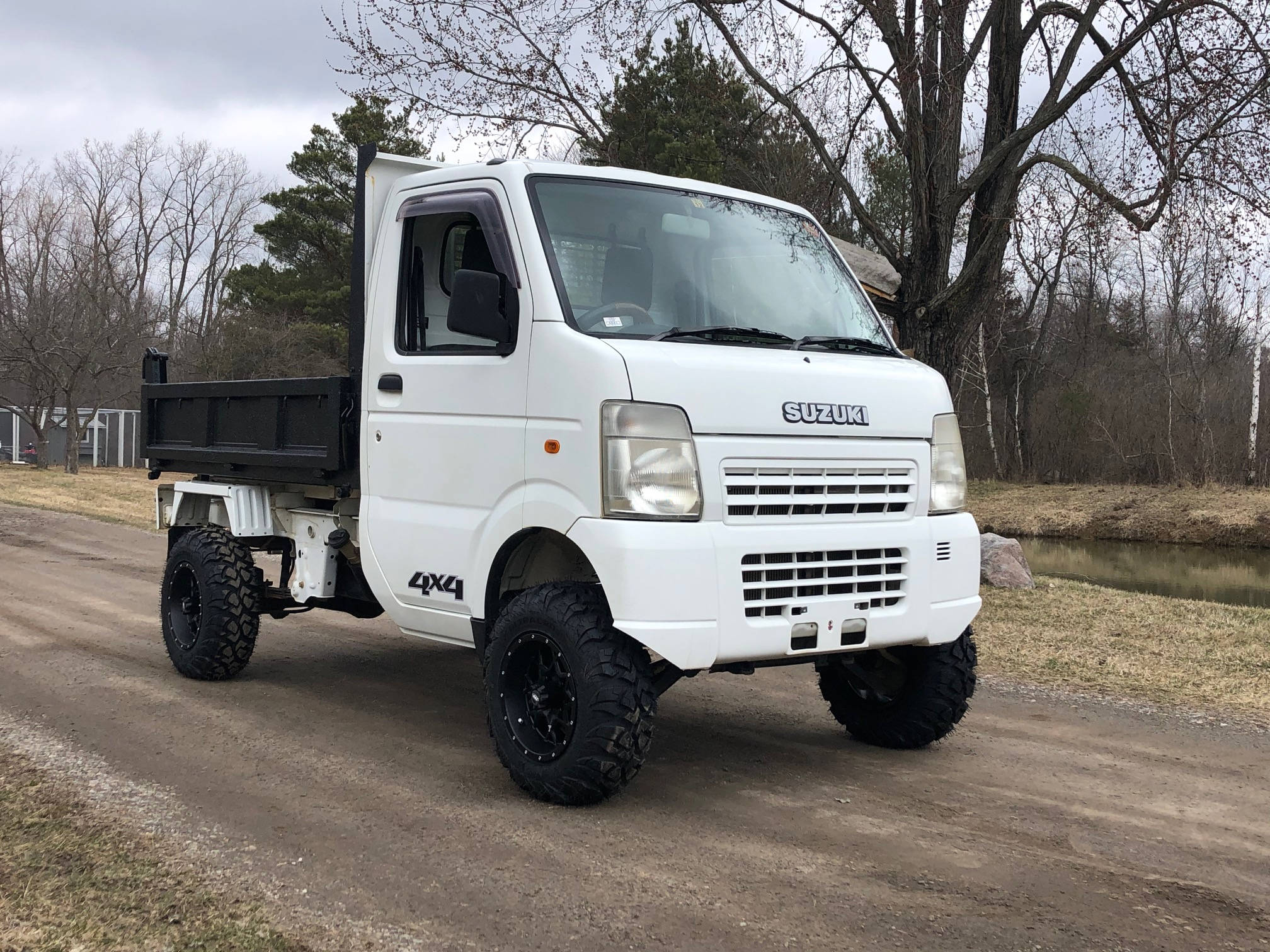 Suzuki 桌布 壁紙 — 一輛白色鈴木4x4自卸車，配備黑色輪胎，停泊在溪流和光禿樹木旁邊的泥土路上。