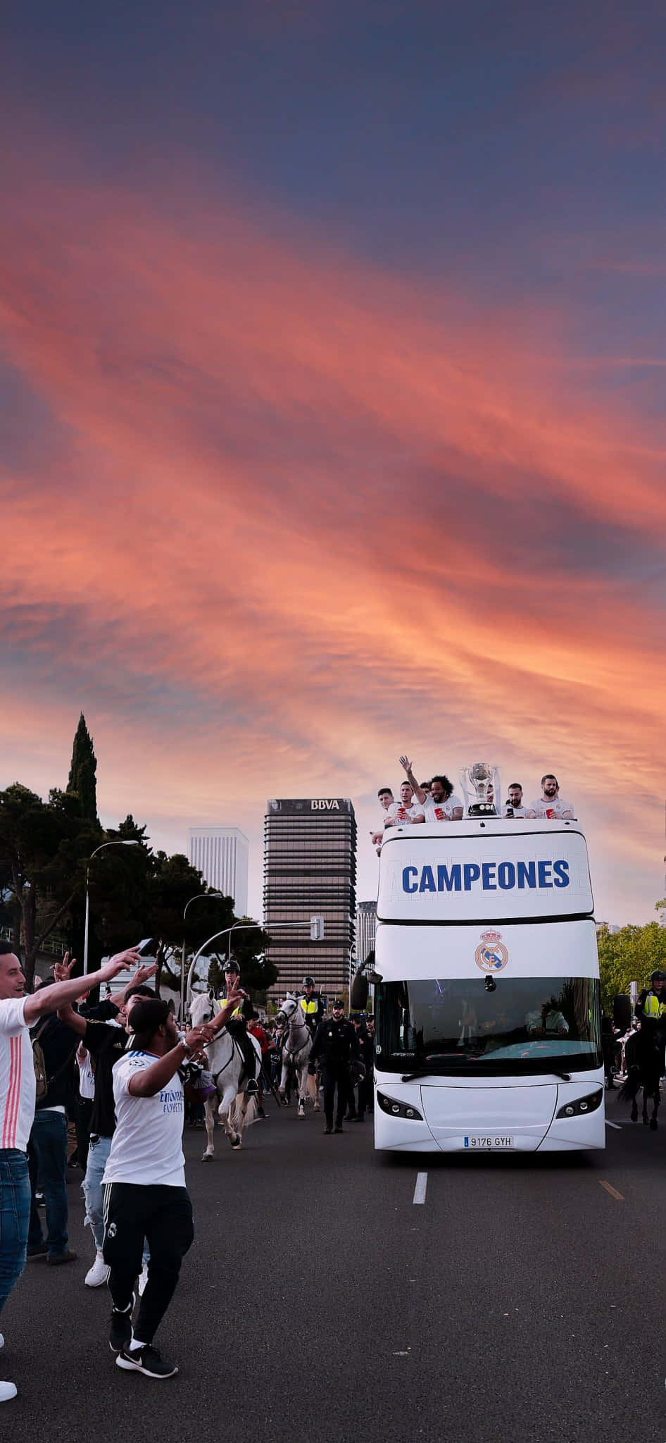 Bus 桌布 壁紙 — 一輛白色雙層巴士，上面標有「CAMPEONES」，載著慶祝的足球運動員穿過城市街道，球迷們在熾熱的日落天空下歡呼。