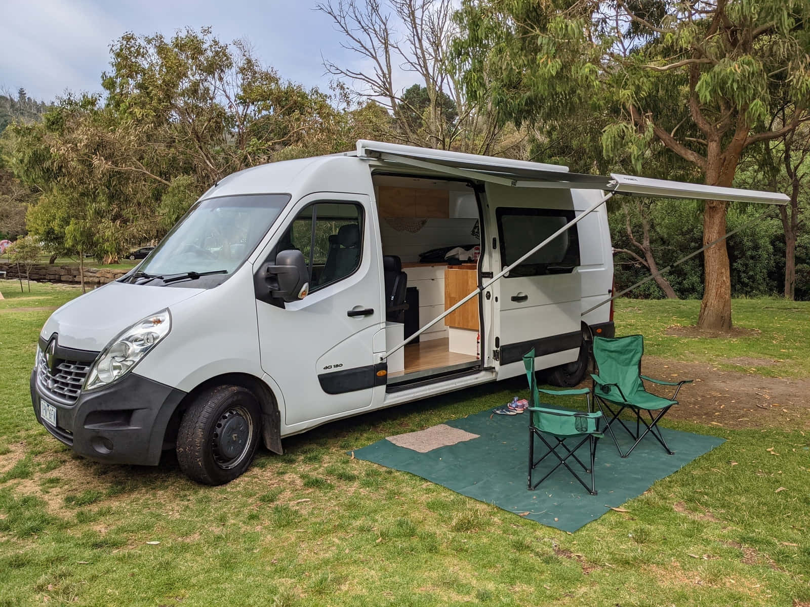Van 桌布 壁紙 — 一輛白色露營車停在草地上，側門打開，遮陽篷展開，露出內部廚房和外面兩張綠色露營椅。