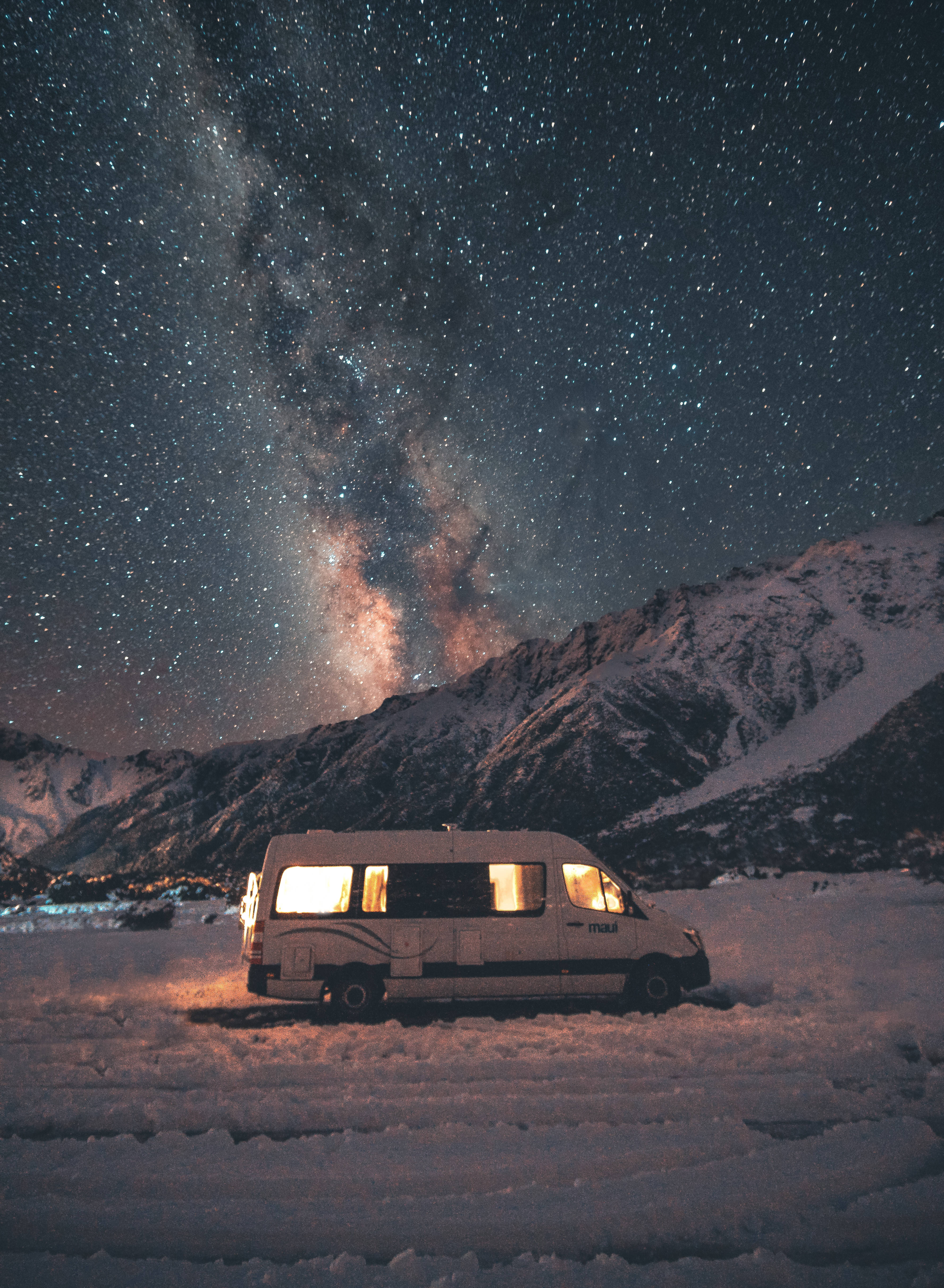 Vans 桌布 壁紙 — 一輛白色露營車在星空下溫暖地發光，銀河在白雪覆蓋的山脈上空彎曲。