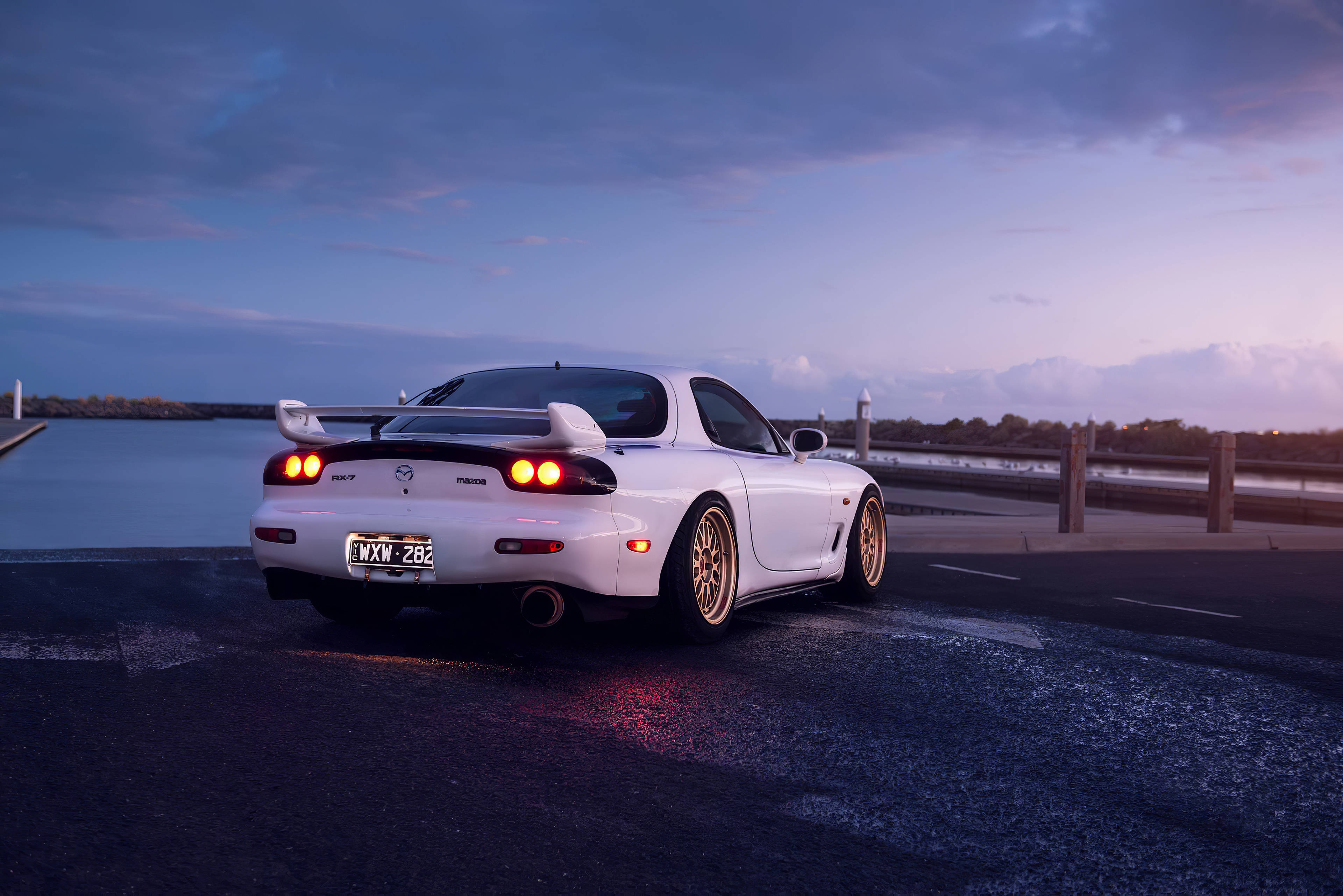 Mazda 桌布 壁紙 — 一輛白色馬自達RX-7跑車配金色輪圈，在紫色天空下，於平靜水邊的濕滑道路上，在黃昏中閃耀。