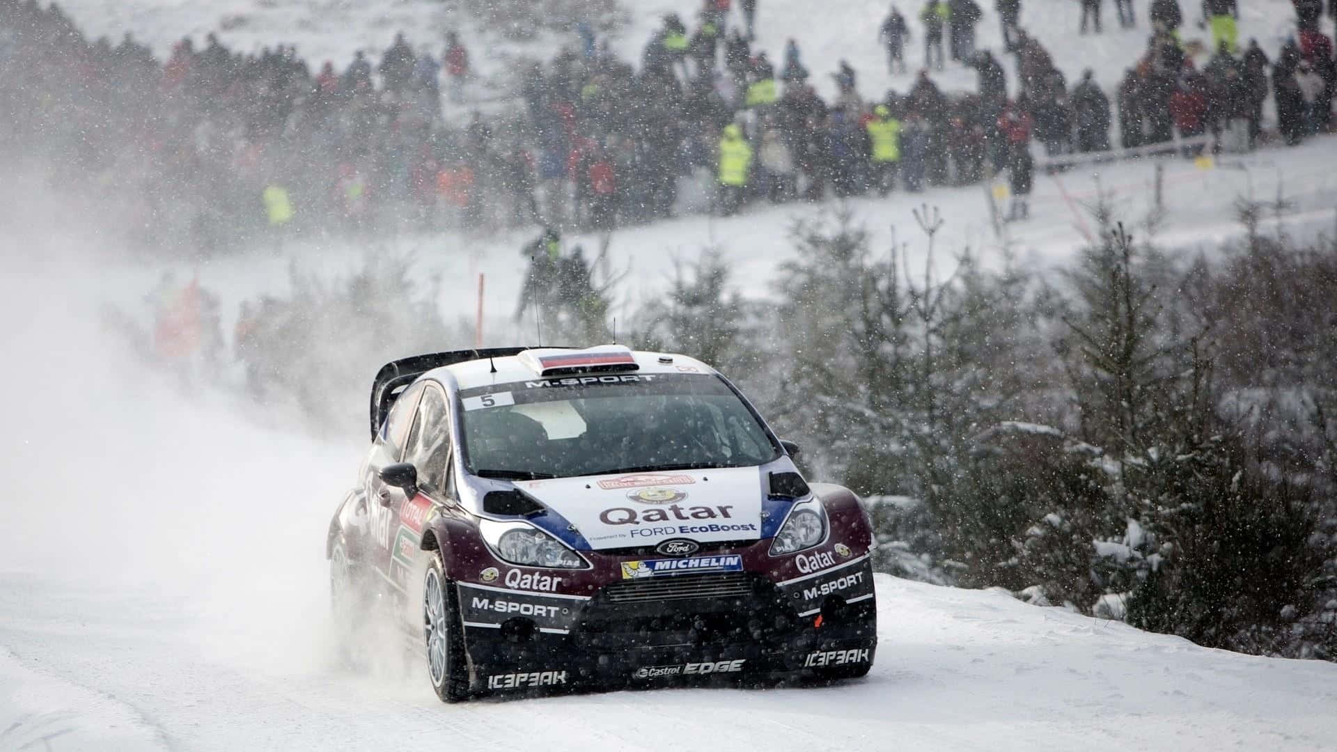 Ice Racing 桌布 壁紙 — 一輛福特拉力賽車在雪地森林賽道上飛馳，揚起雪花，觀眾在旁觀看。