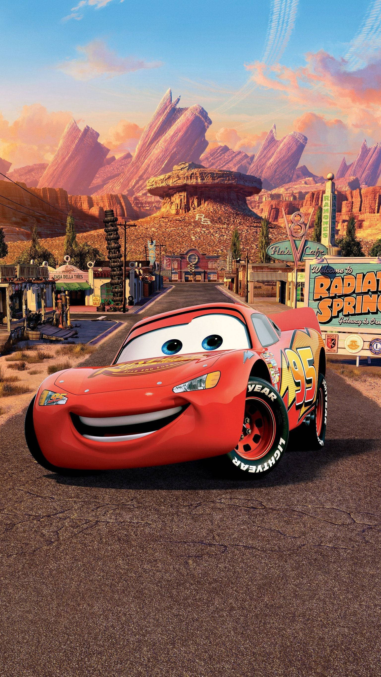 Lightning Mcqueen 桌布 壁紙 — 一輛笑容滿面的紅色賽車，在溫暖的日落天空下，自豪地站在一個有岩石山脈的沙漠小鎮前面。