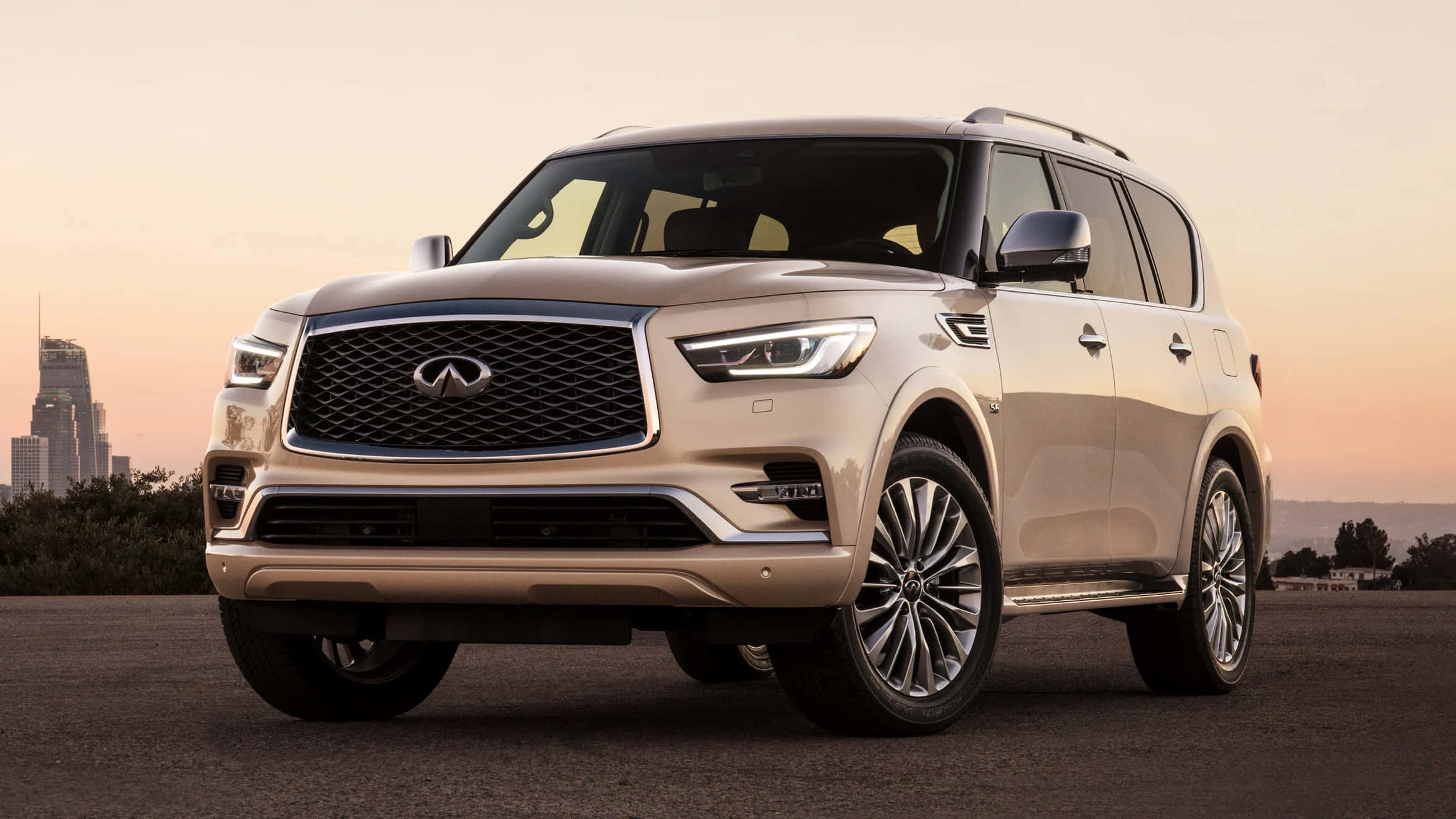 Infiniti Qx80 桌布 壁紙 — 一輛米色Infiniti QX80 SUV停泊在瀝青路上，背景是城市天際線和日落。