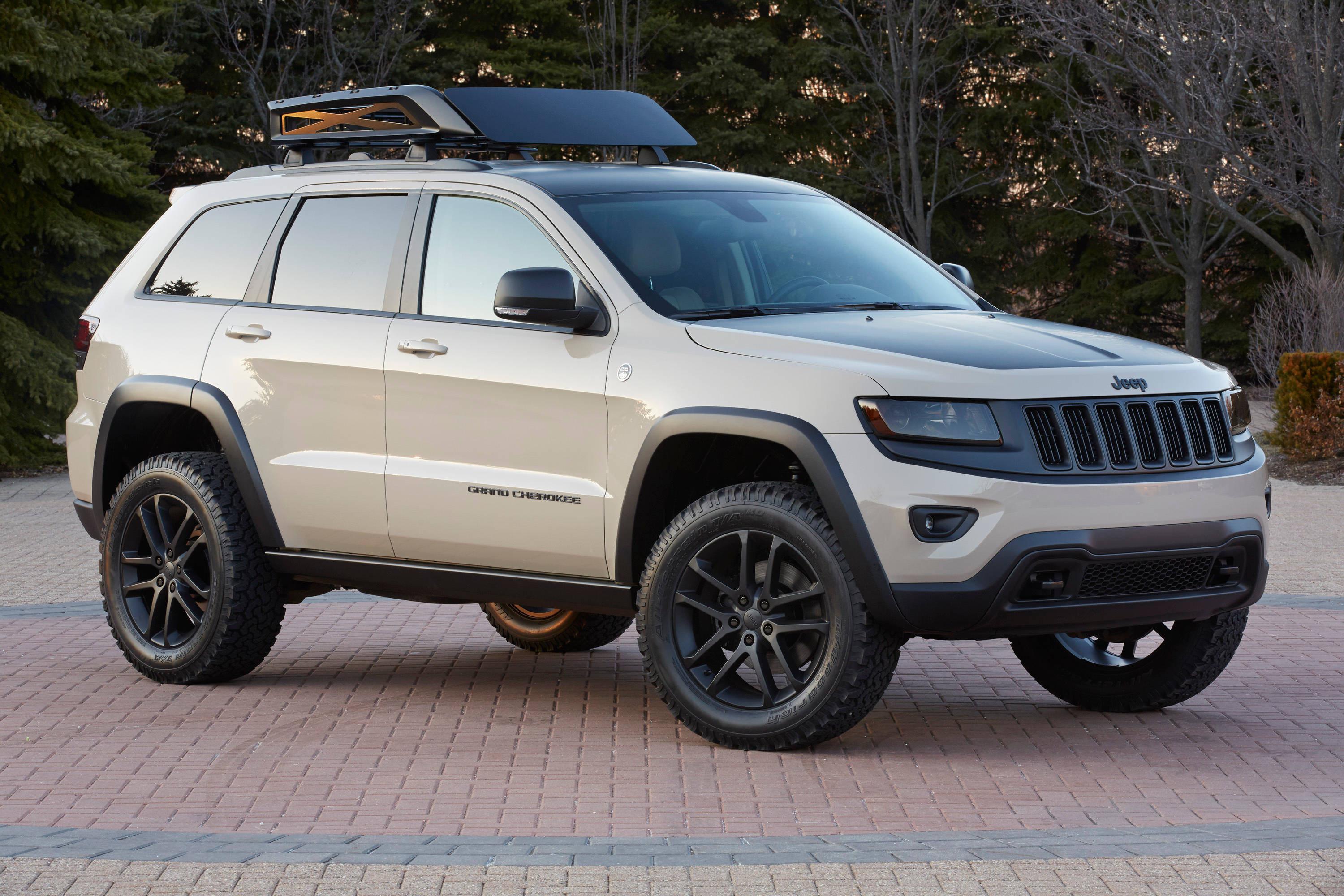 吉普 桌布 壁紙 — 一輛米色Jeep Grand Cherokee，帶有黑色飾邊和車頂行李架，停在磚砌車道上，周圍樹木環繞。