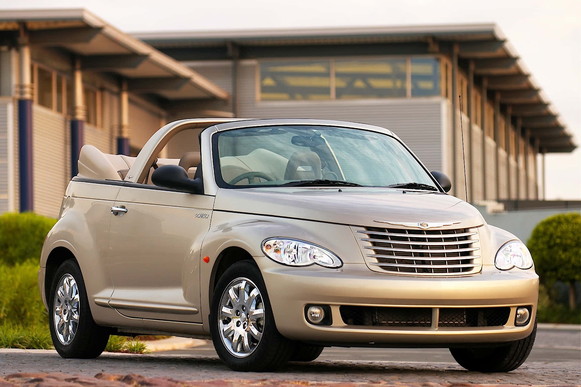 Chrysler Pt Cruiser 桌布 壁紙 — 一輛米色克萊斯勒 PT Cruiser 敞篷車停放在磚砌車道上，背景是現代建築和綠色灌木。