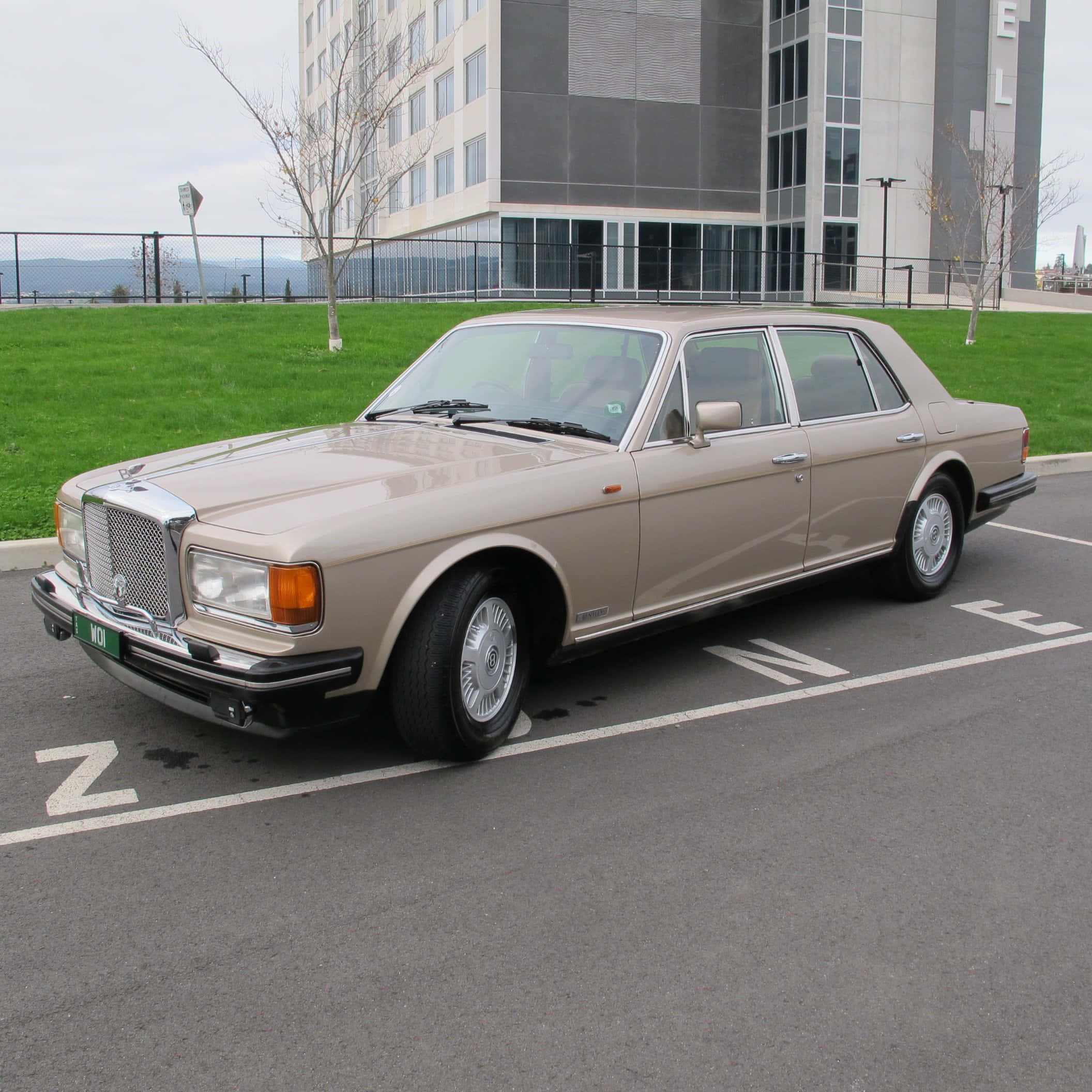 Bentley Eight 桌布 壁紙 — 一輛米色勞斯萊斯銀靈停放在現代建築旁邊的標記車位，建築物周圍有綠色草地和光禿禿的樹木，天空多雲。