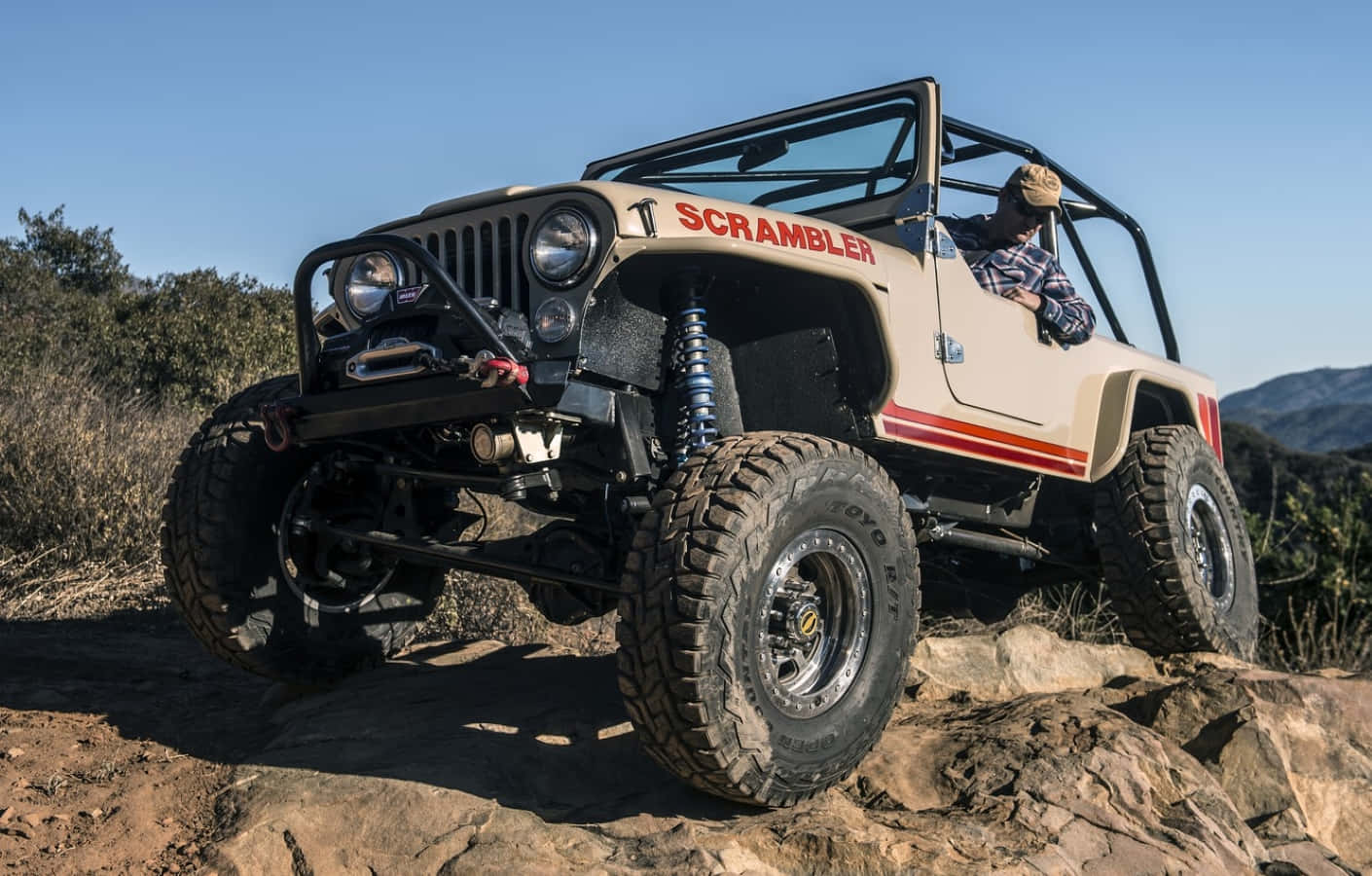 Jeep Cj 桌布 壁紙 — 一輛米色帶紅色條紋的Jeep Scrambler在晴朗藍天下攀爬崎嶇岩石，駕駛員在開放式駕駛艙中。