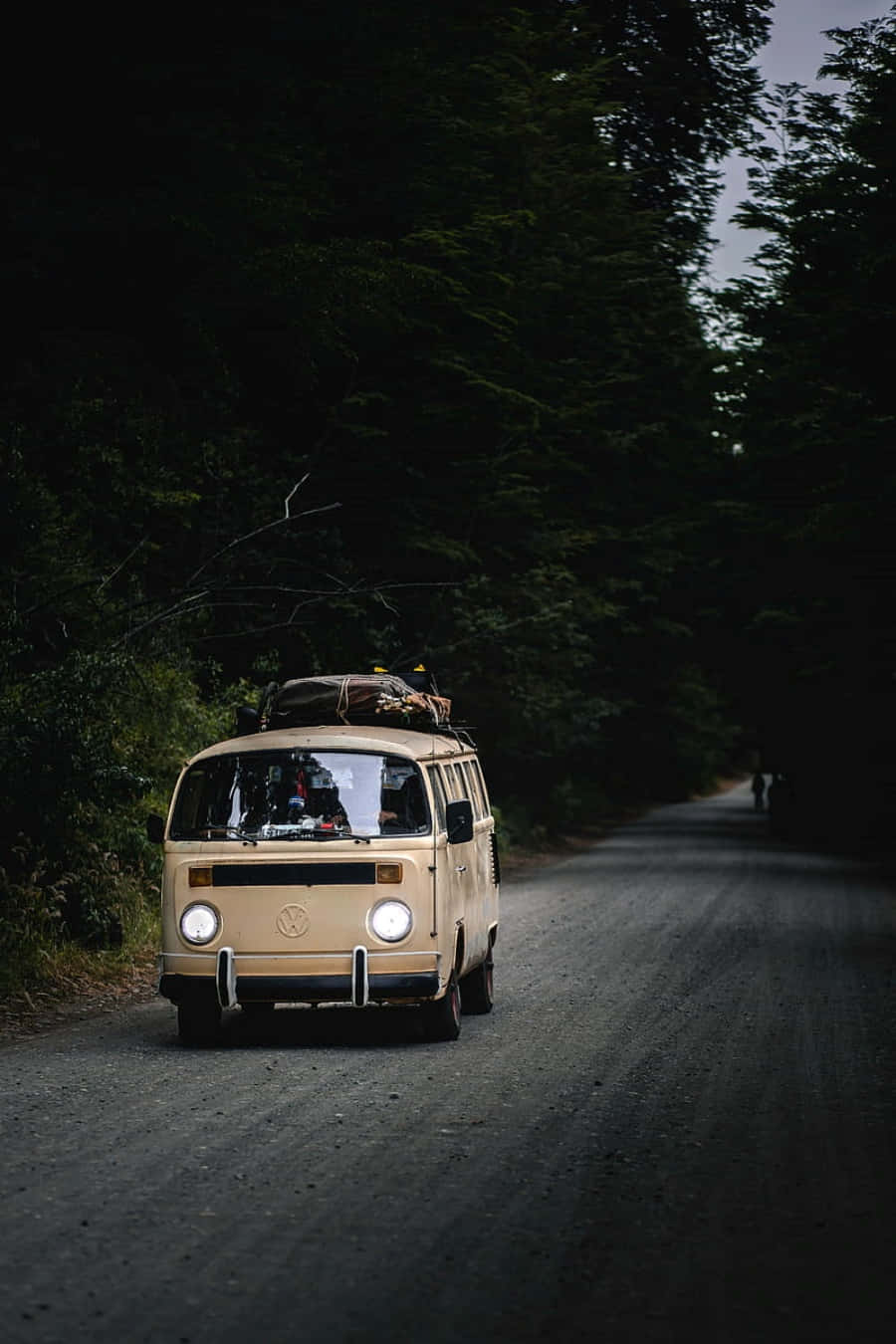 Van 桌布 壁紙 — 一輛米色復古福斯露營車車頂載著行李，在陰鬱的暮色下行駛在一條漆黑的森林道路上。