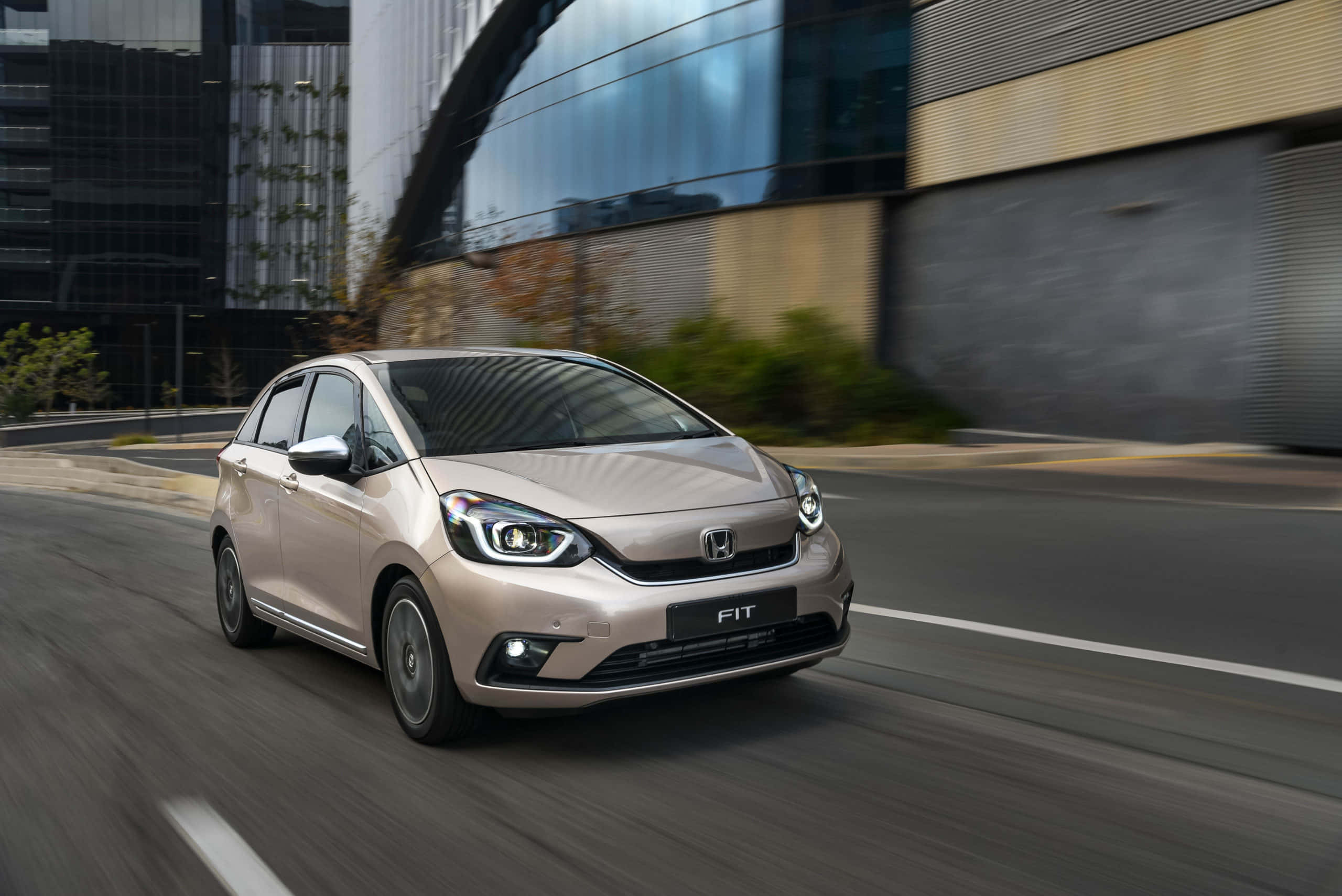 Honda Fit 桌布 壁紙 — 一輛米色本田飛度在模糊的現代建築和綠化背景下的城市街道上飛馳。