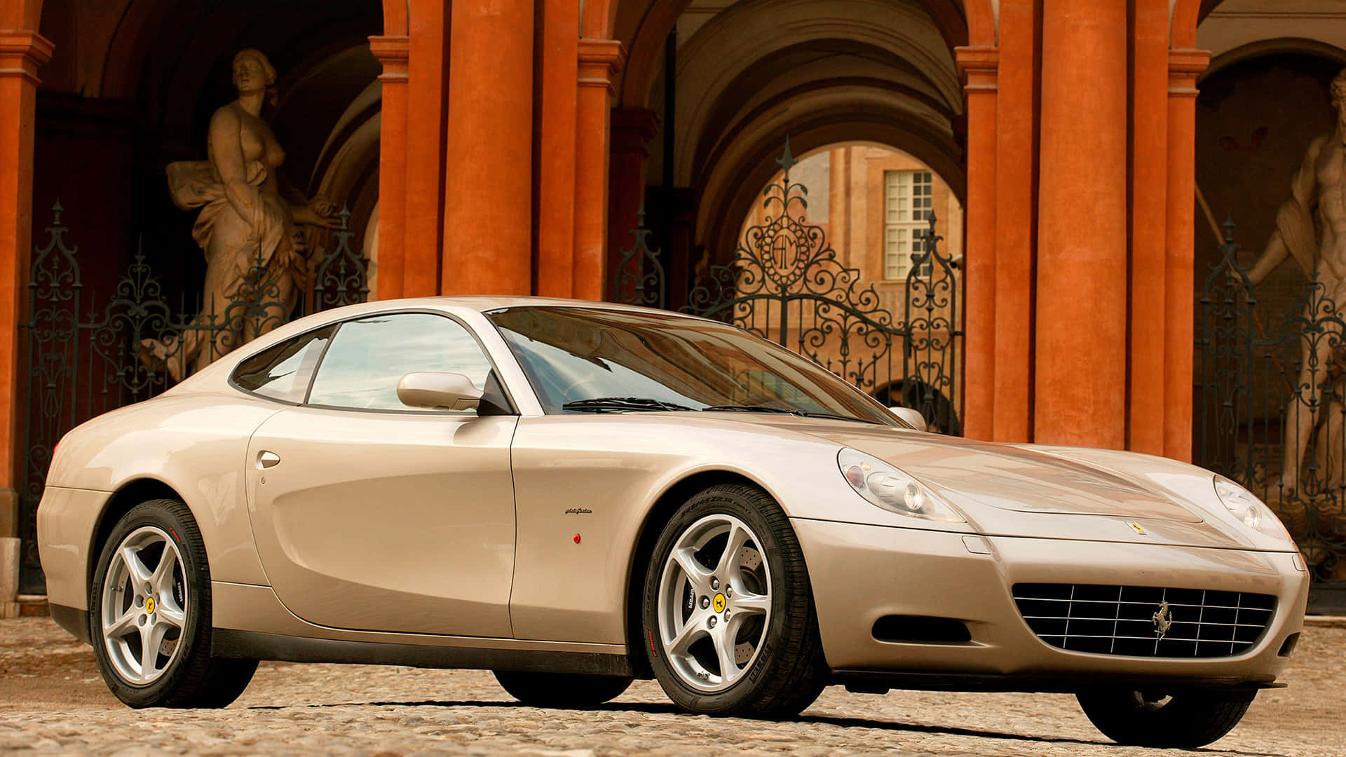 Ferrari 612 Scaglietti 桌布 壁紙 — 一輛米色法拉利612 Superamerica停泊在鵝卵石路上，前方是帶有雕像的華麗橙色石拱門。