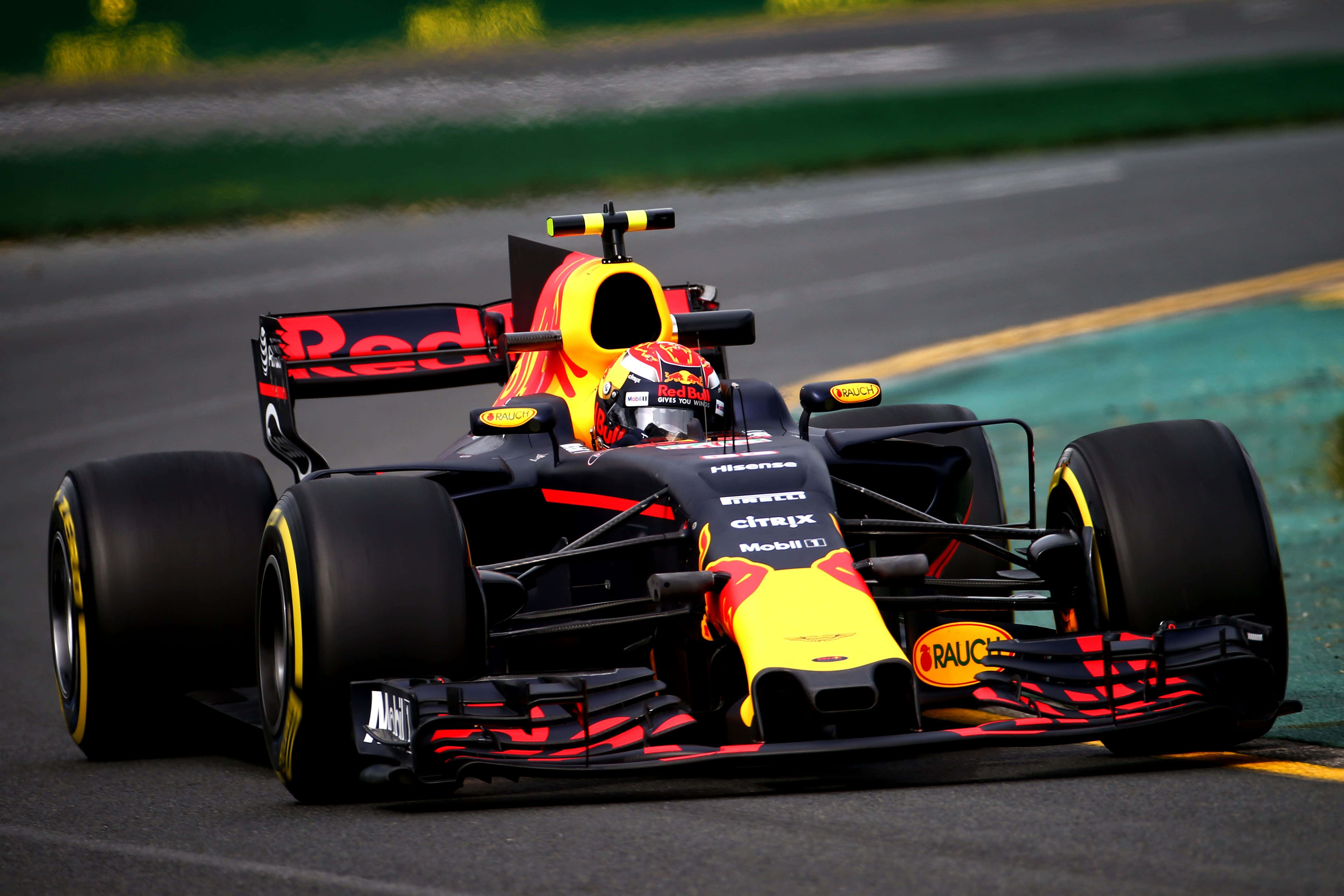 Max Verstappen 桌布 壁紙 — 一輛紅牛一級方程式賽車在賽道上疾馳，帶有鮮豔的黃色和紅色點綴，背景是模糊的綠色和灰色。