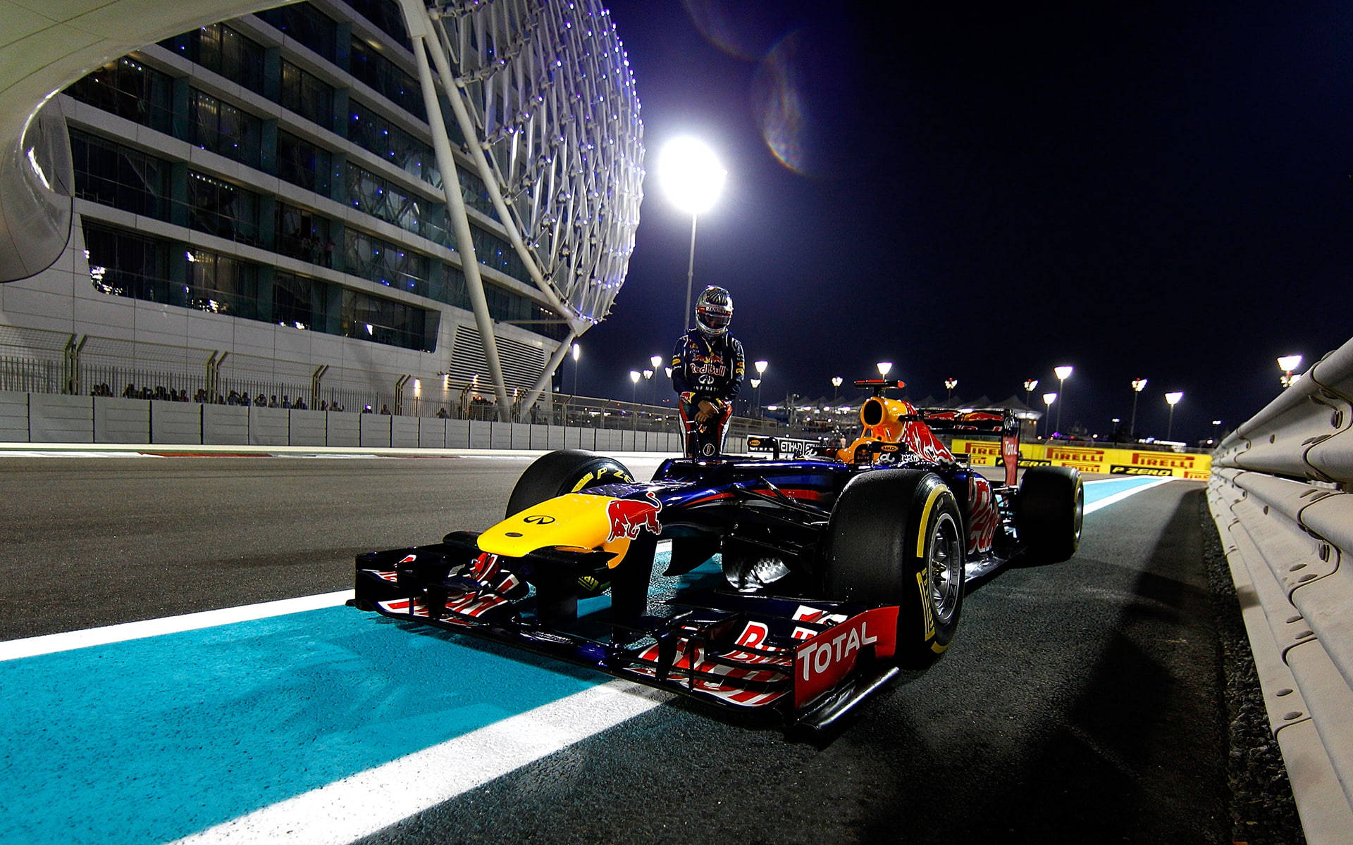 F1 桌布 壁紙 — 一輛紅牛一級方程式賽車與車手在夜間準備就緒，停在明亮照明的賽道上，旁邊是一座未來主義的體育場。